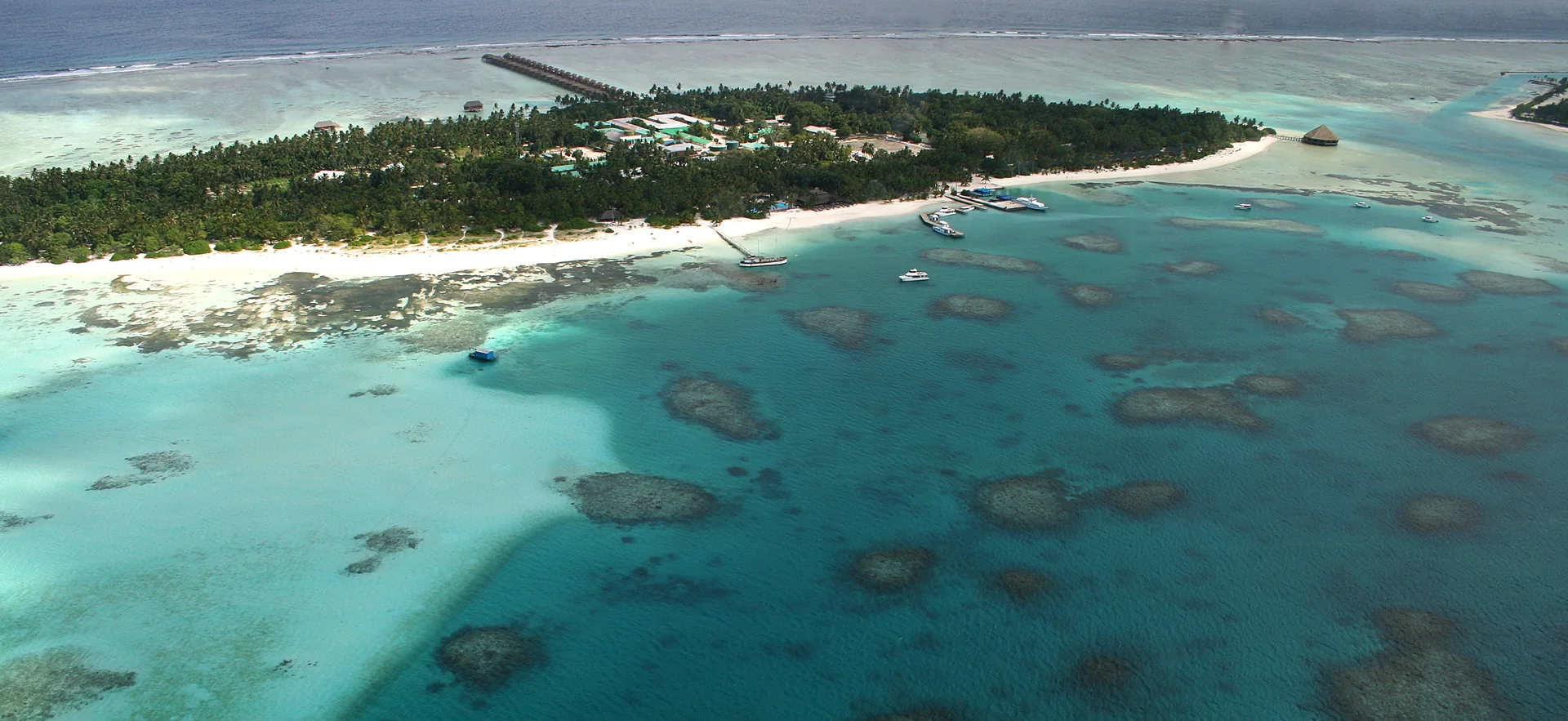 Vista aérea ampla de uma ilha-resort nas Maldivas, com vegetação, faixa de areia e construções pequenas em meio à lagoa, sem hóspedes identificáveis.
