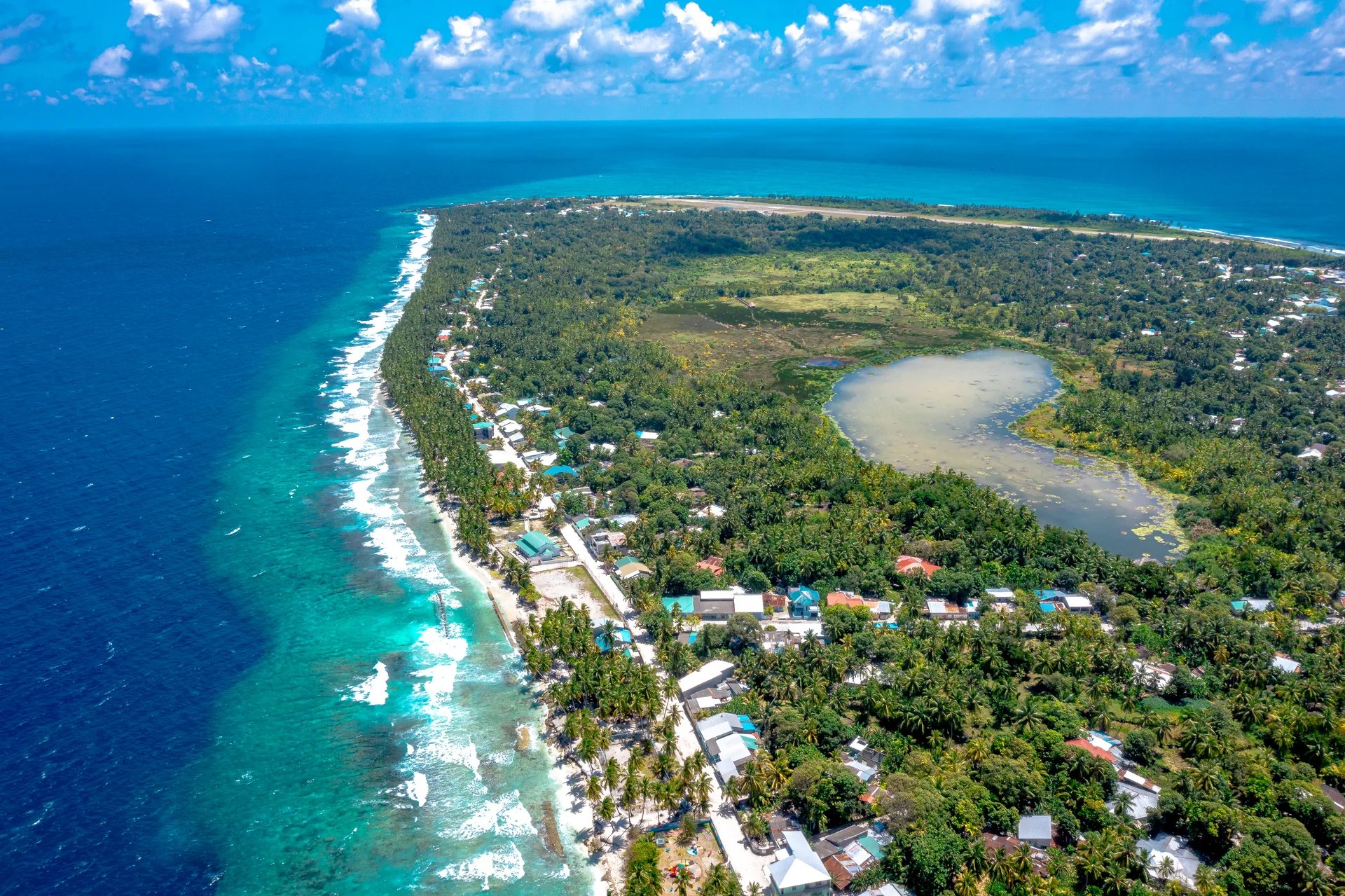 Vista aérea ensolarada de Fuvahmulah, ilha habitada das Maldivas, com costa, casas, vegetação e lago interior, sem pessoas identificáveis.