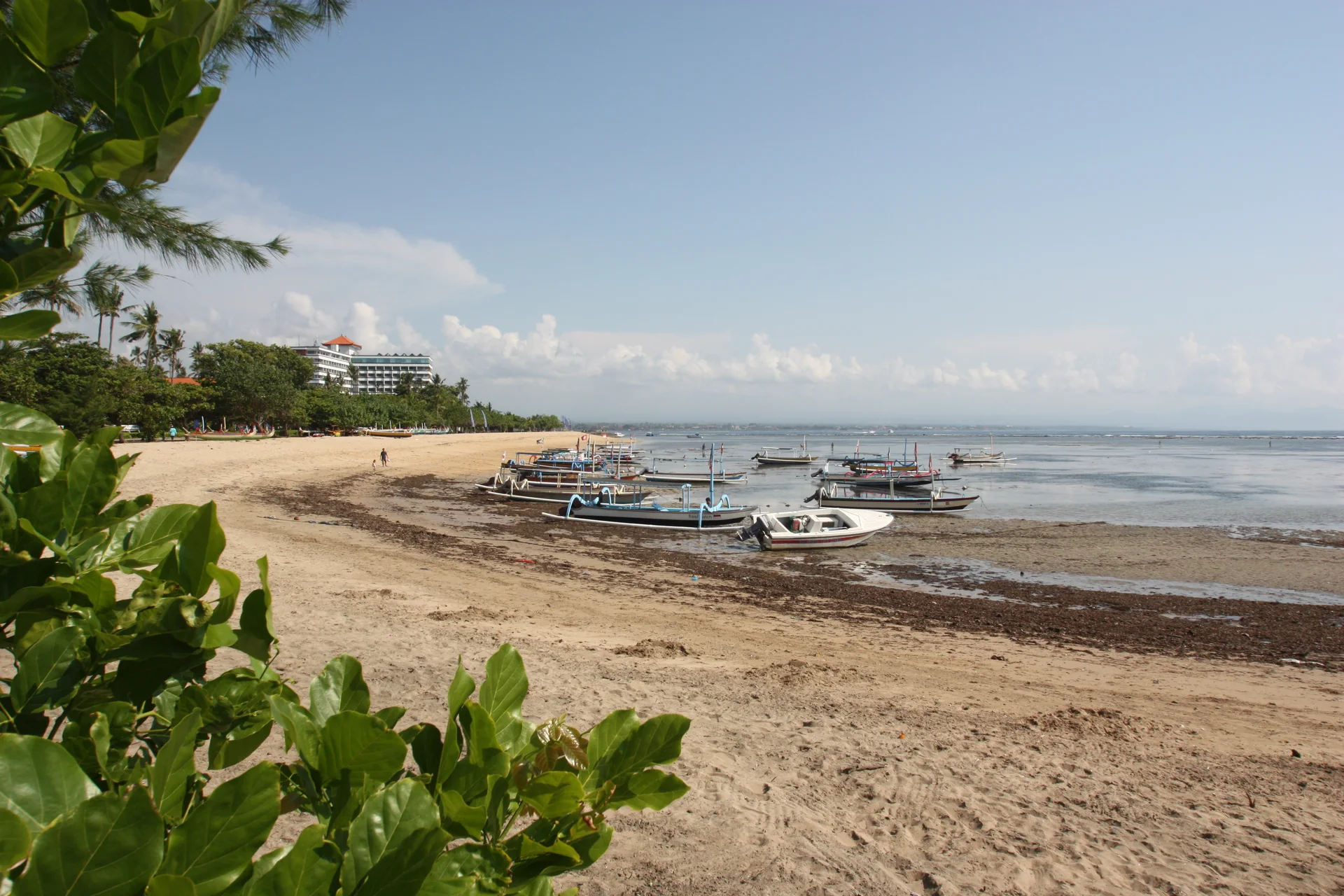 Faixa de areia de Sanur com barcos pequenos junto à água, vegetação e construções ao fundo sob céu claro.