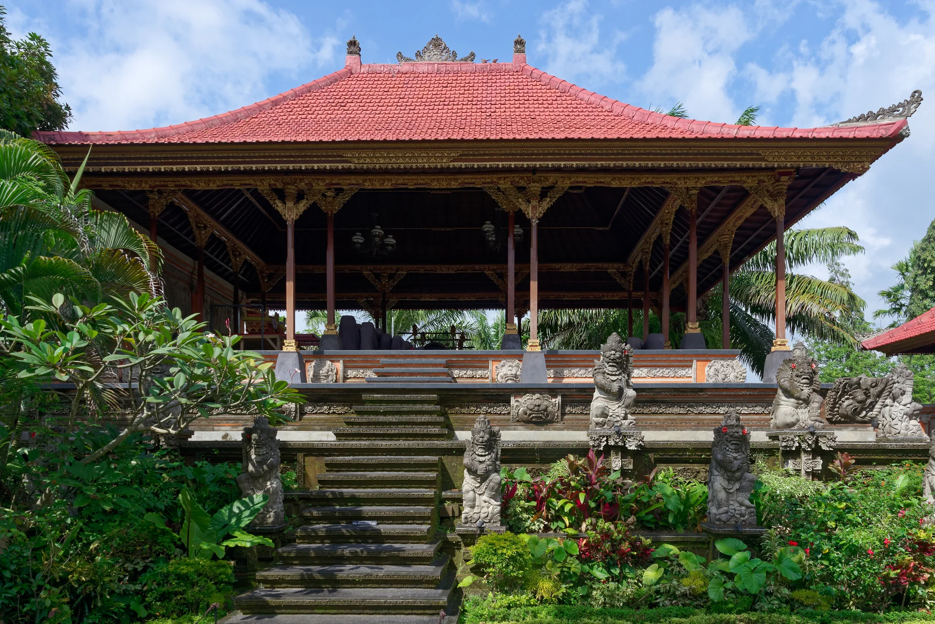 Pavilhão de telhado vermelho e ornamentos esculpidos no Palácio de Ubud, em meio a jardim tropical.