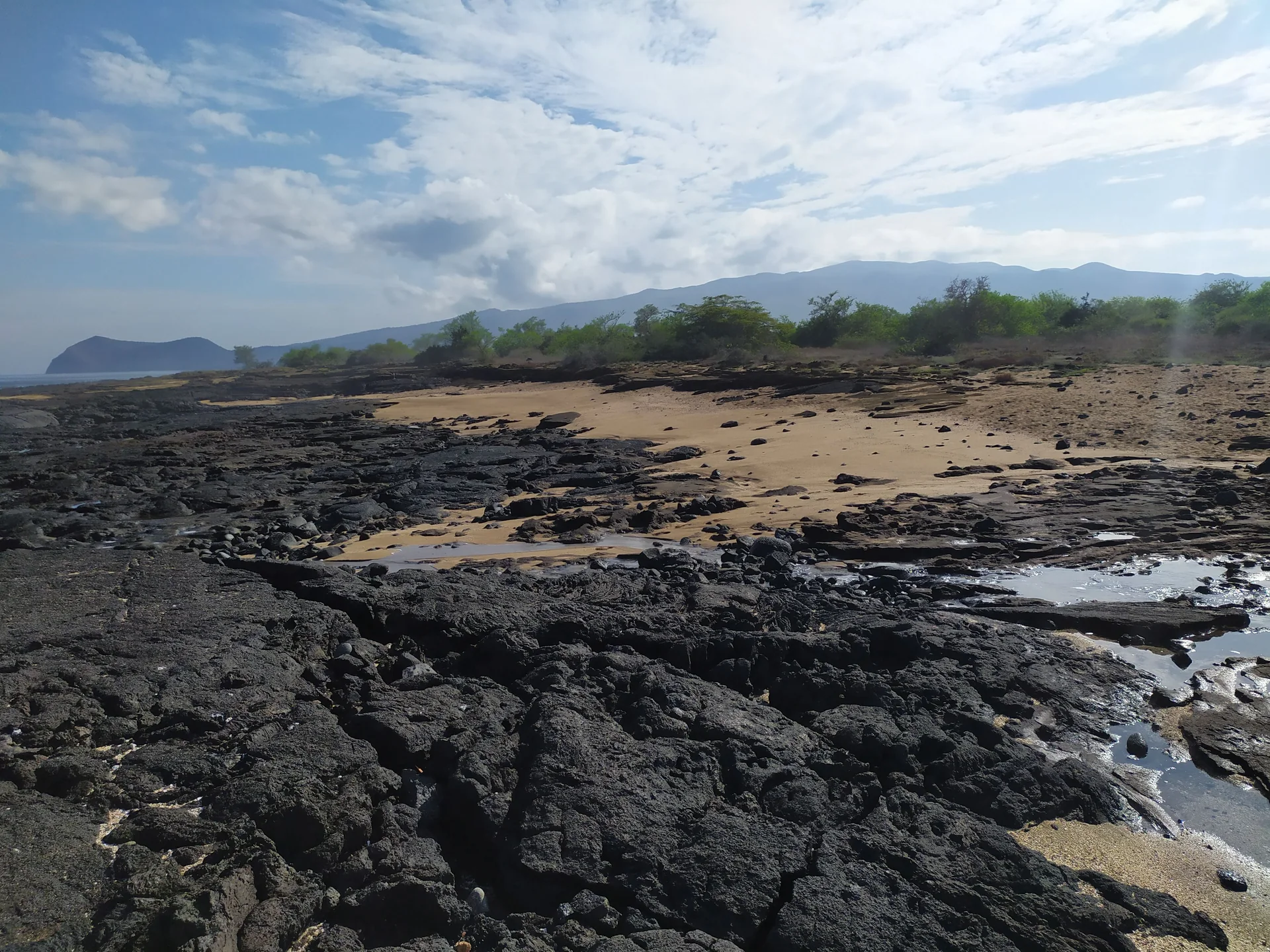 Faixa de areia entre formações de lava escura, vegetação costeira e relevo ao fundo em Galápagos, sem animais ou pessoas.