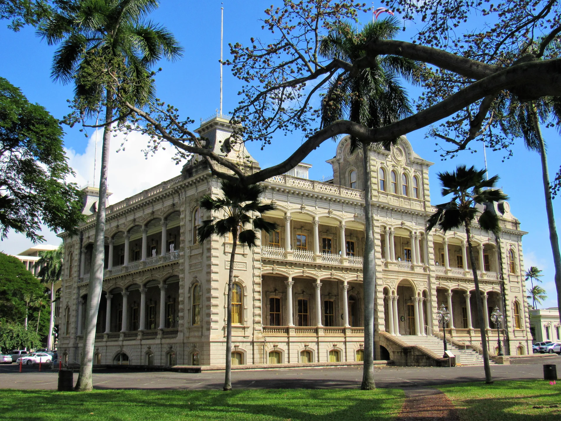 Fachada externa do ʻIolani Palace, em Honolulu, enquadrada por gramado, palmeiras e árvores.