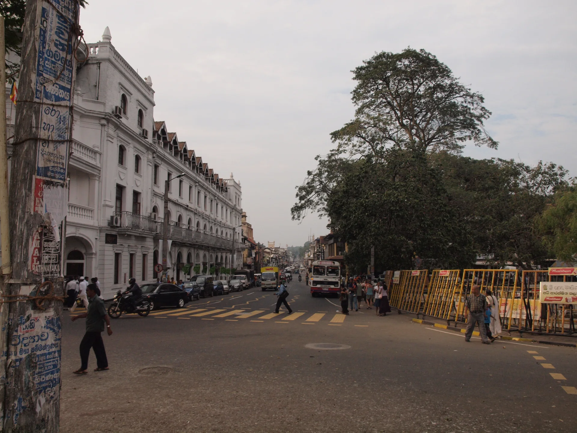 Rua urbana de Kandy com pessoas, veículos, edifícios e encosta arborizada ao fundo.
