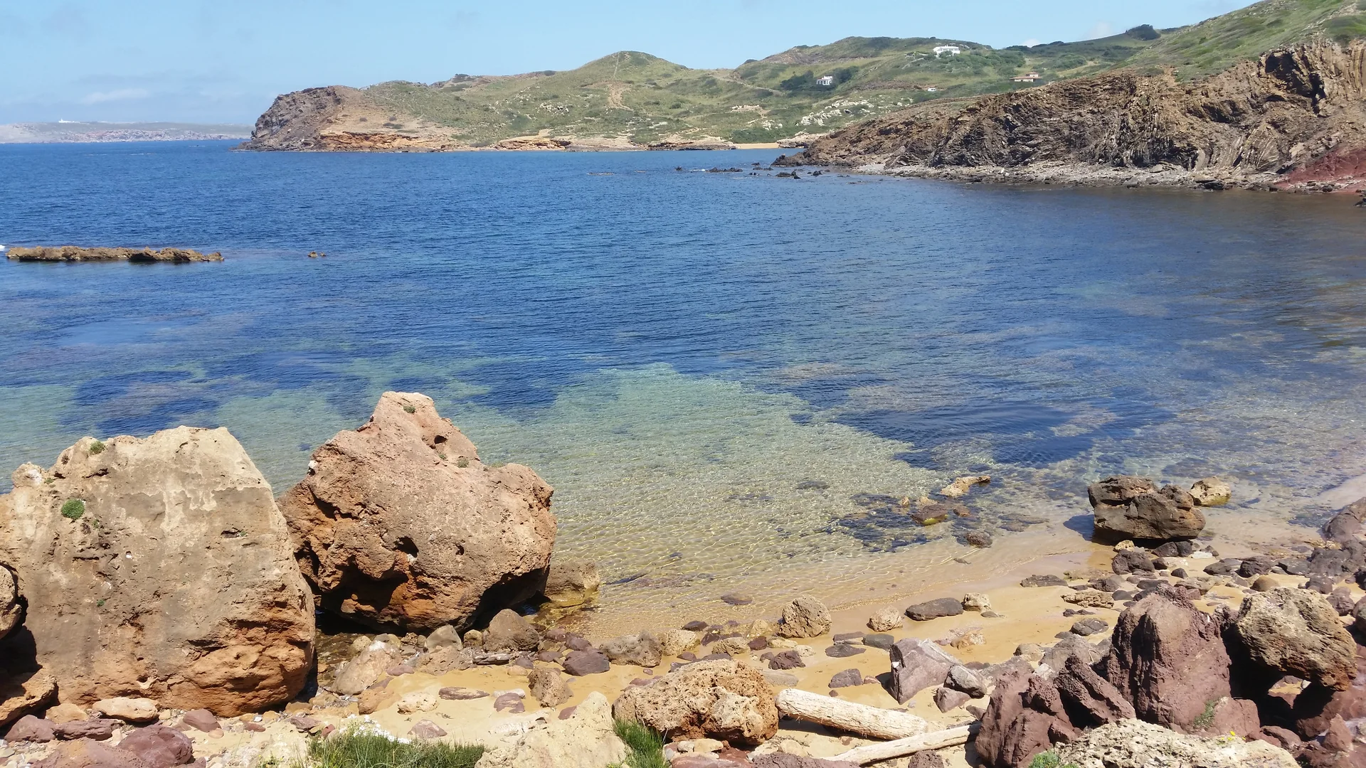 Enseada rochosa na área de Cavalleria, no norte de Menorca, com água azul, falésias e vegetação baixa sob luz clara.
