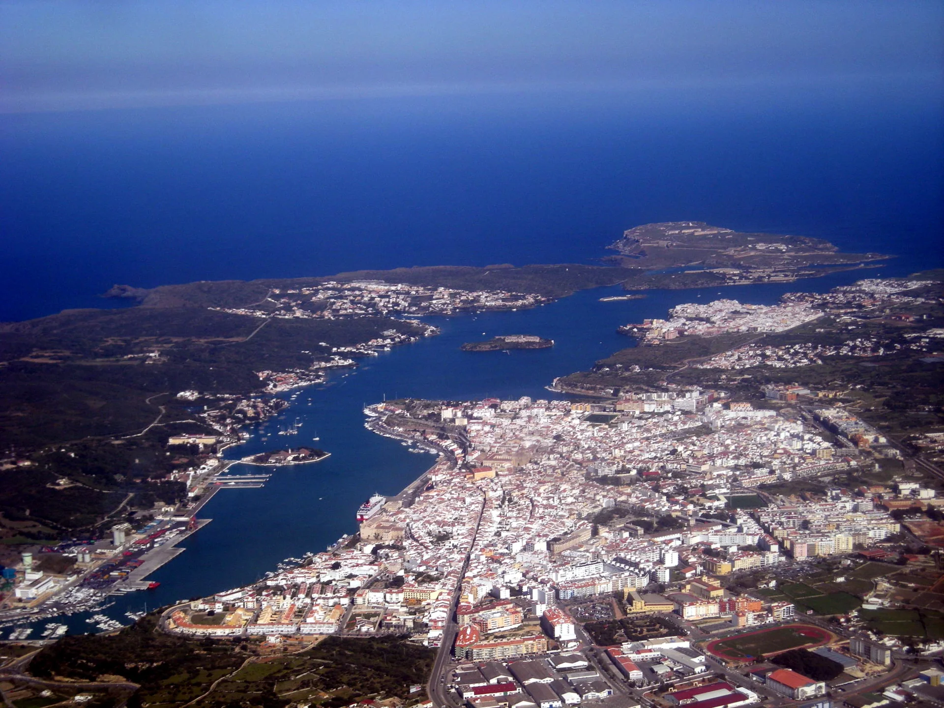 Vista aérea ampla de Maó e de seu porto, com o núcleo urbano, a enseada e a paisagem costeira ao redor.