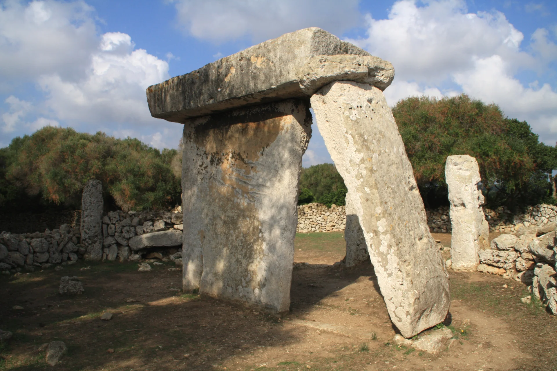Taula de pedra no assentamento talayótico de Talatí de Dalt, enquadrada por vegetação e muro de pedra em Menorca.