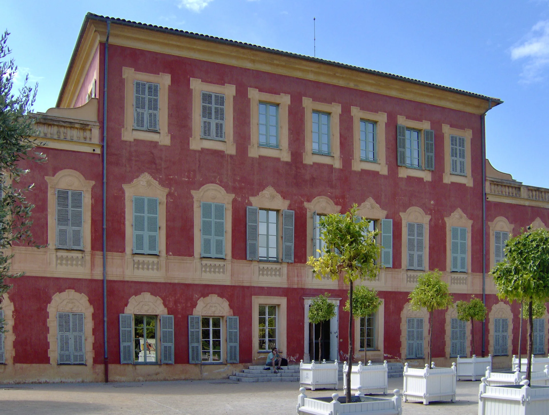 Fachada do Musée Matisse em Cimiez, em Nice, com paredes vermelhas, persianas e árvores no primeiro plano.