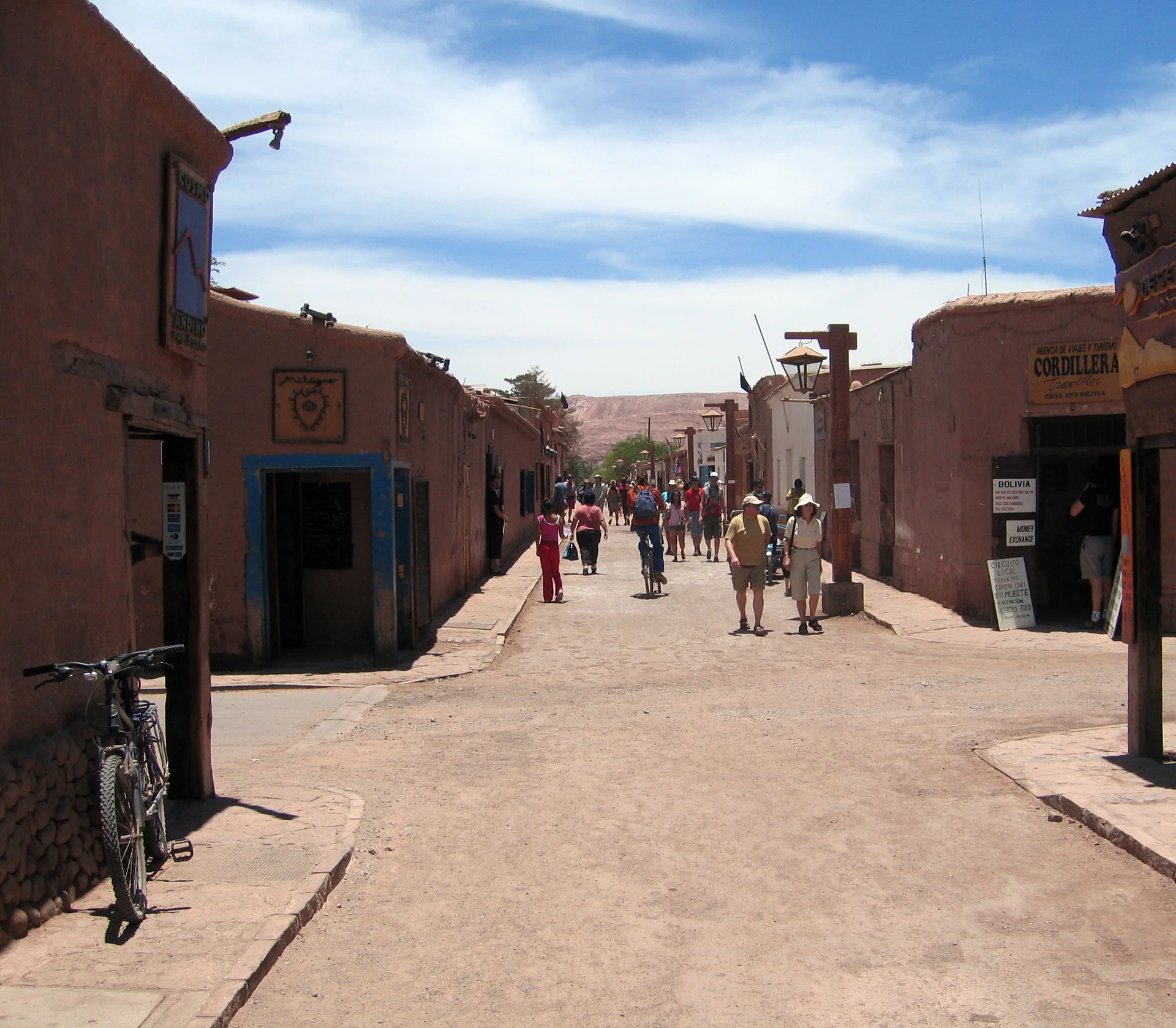 Rua movimentada de San Pedro de Atacama, com fachadas de adobe, bicicletas e visitantes sob céu claro.