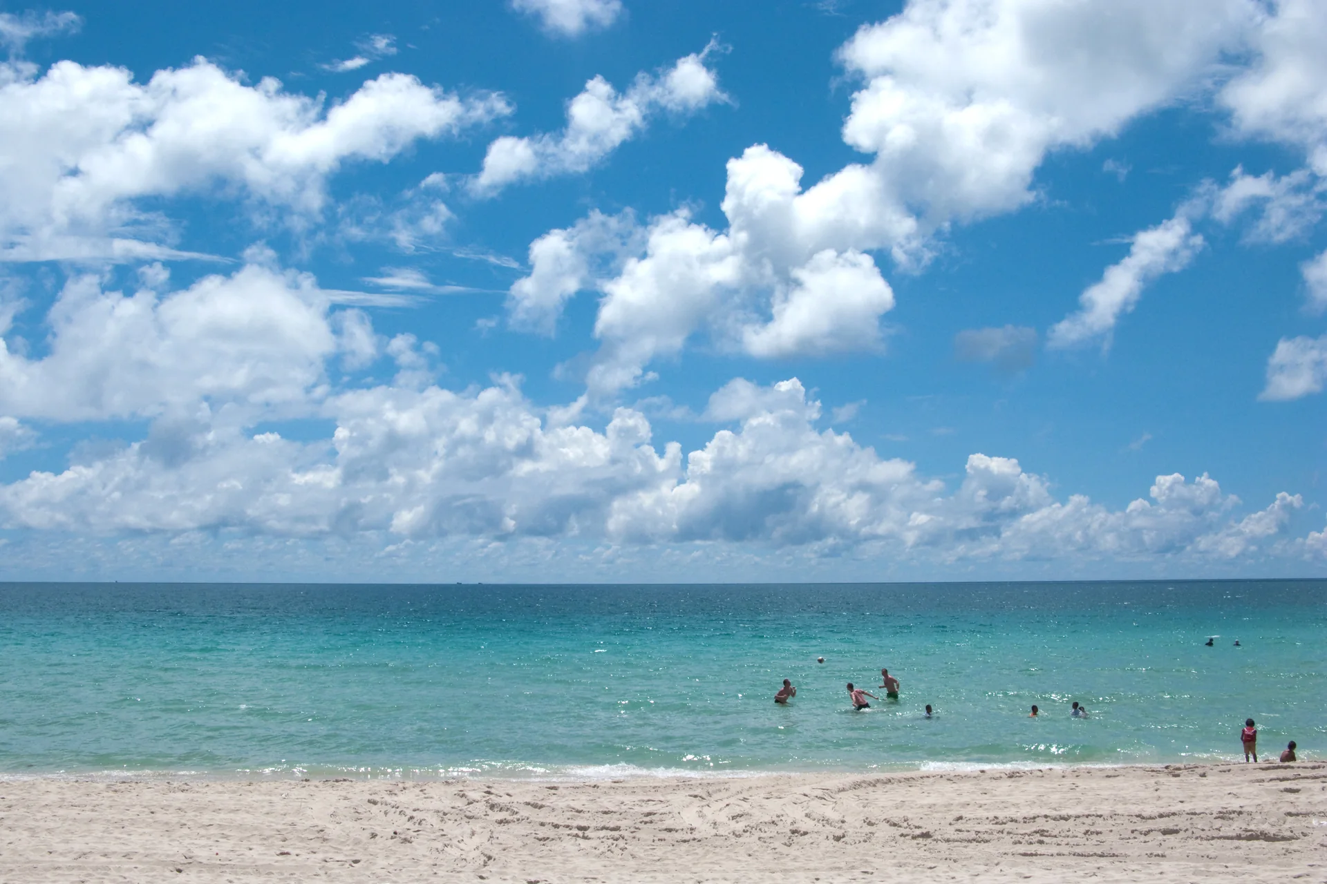 Faixa de areia em Koh Samui, com mar azul-turquesa, banhistas ao longe e céu azul com nuvens durante o dia.