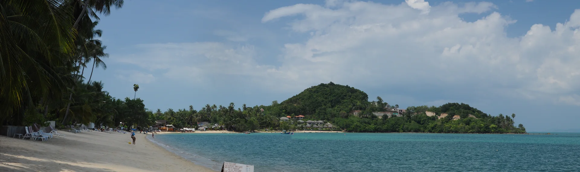 Panorama de praia em Koh Samui, com areia, coqueiros, vegetação costeira e mar calmo durante o dia.