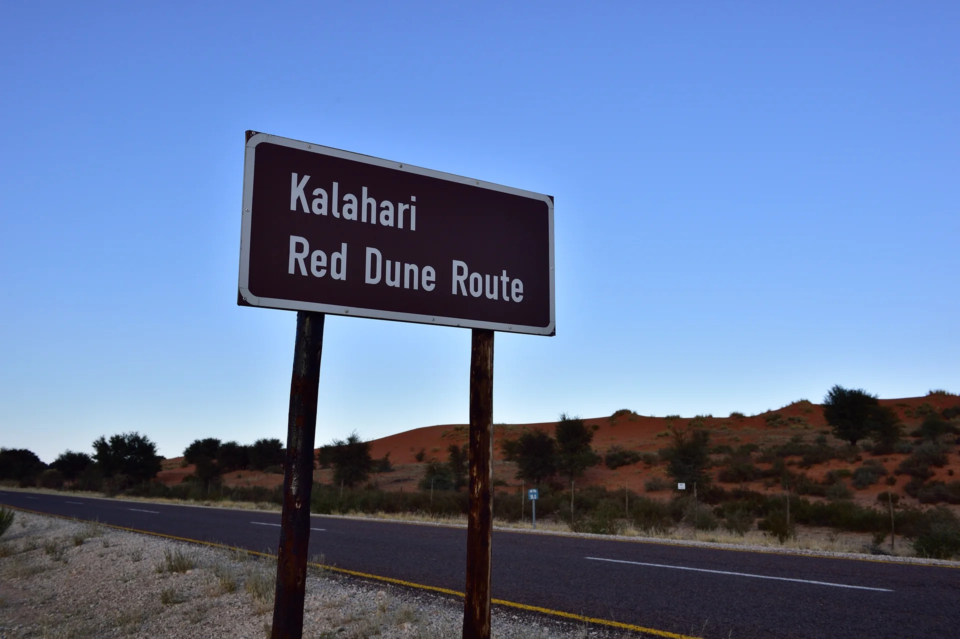 Placa da Red Dune Route junto a estrada e dunas avermelhadas no Parque Transfronteiriço Kgalagadi, sob céu azul claro.