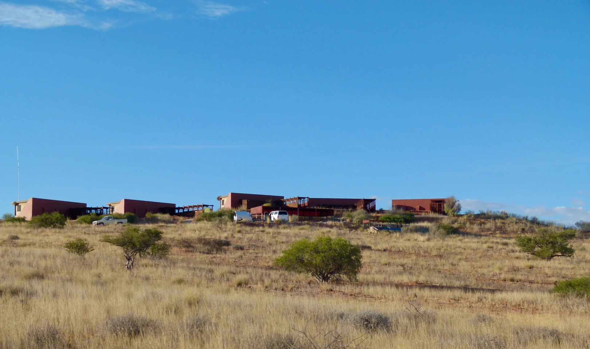 Cabanas baixas do Kieliekrankie Wilderness Camp sobre terreno árido do Kgalagadi, sob céu azul intenso.