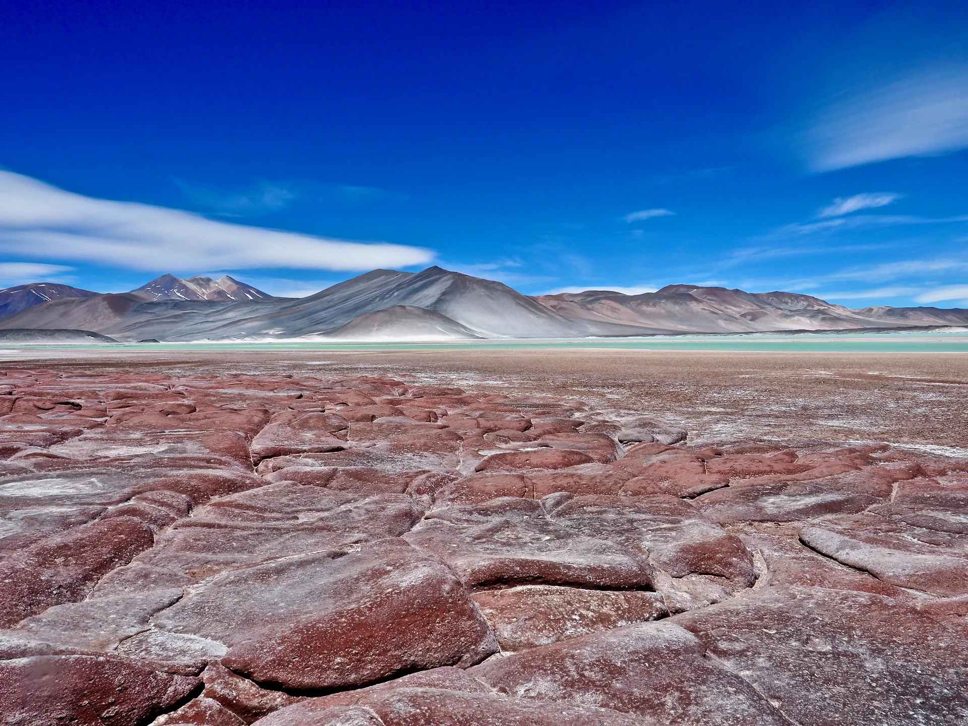 Rochas avermelhadas em primeiro plano diante de águas rasas e montanhas do deserto do Atacama sob céu azul.