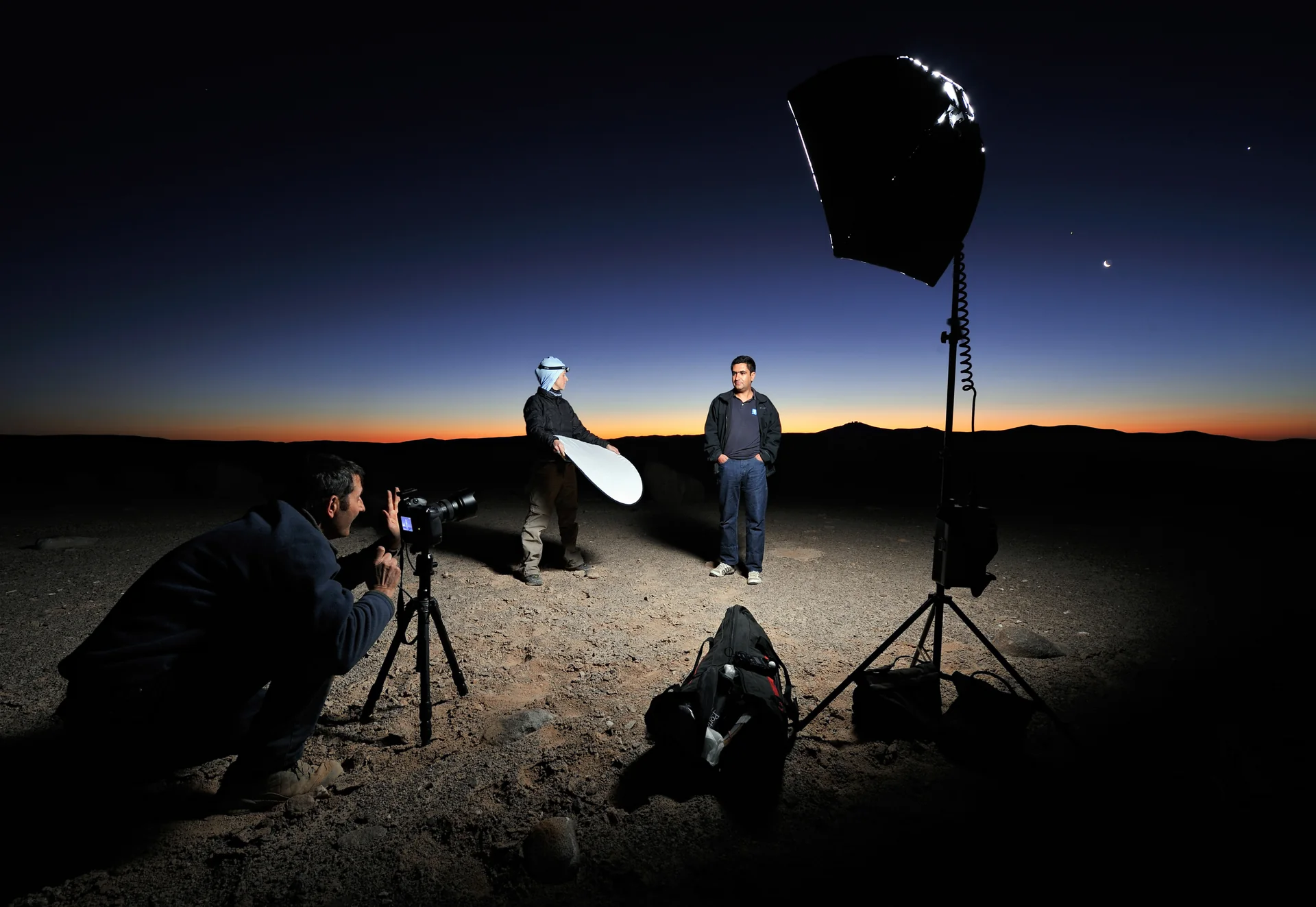 Equipe em atividade noturna no Deserto do Atacama, com iluminação controlada, silhuetas de montanhas e Lua crescente.