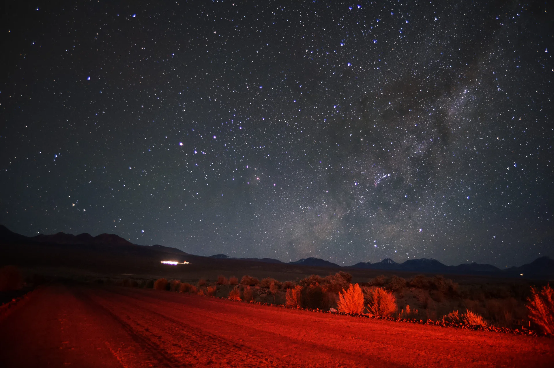 Céu estrelado sobre montanhas e vegetação baixa no Deserto do Atacama, com uma luz vermelha fraca iluminando o primeiro plano.