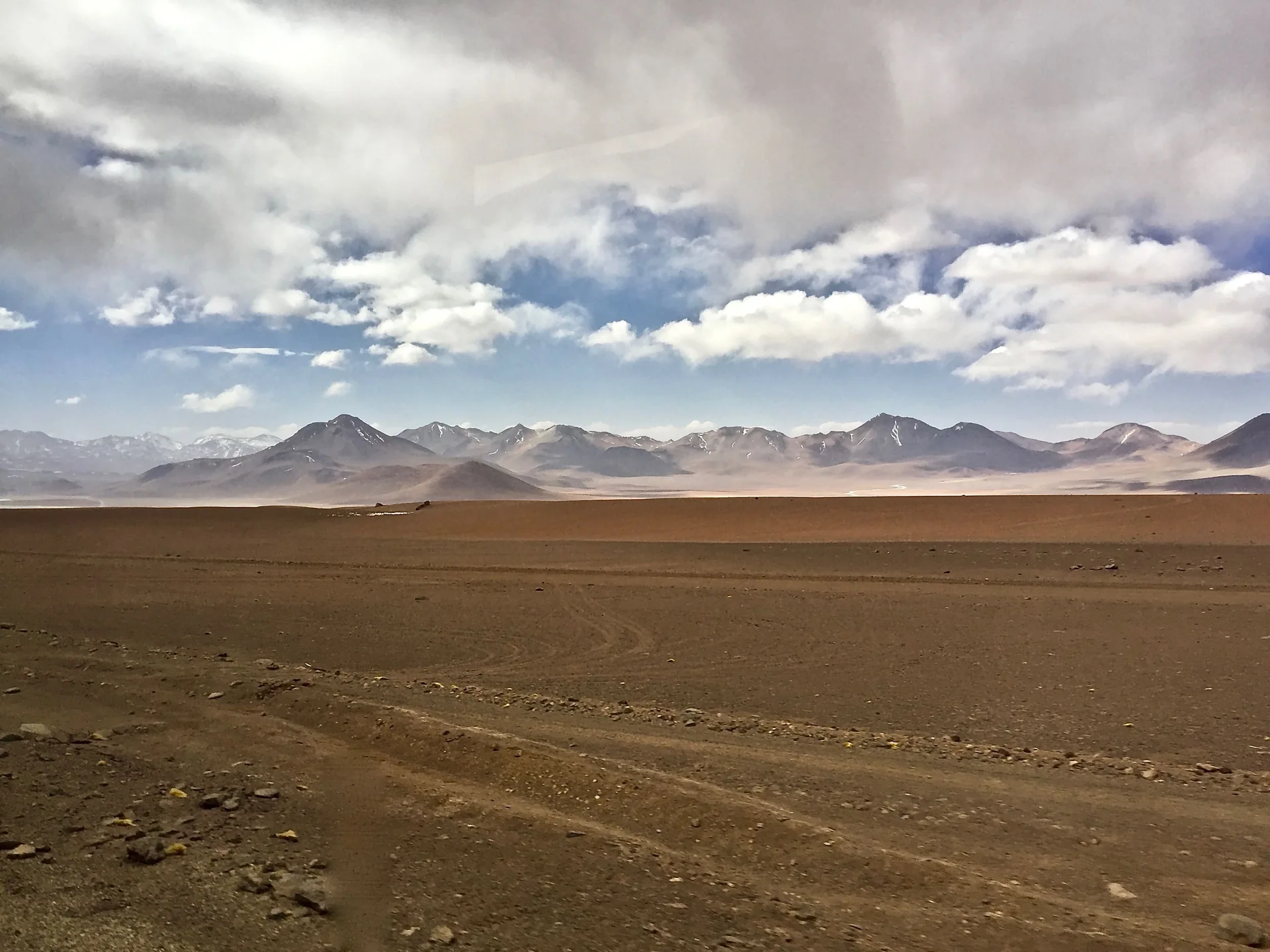Planície árida do altiplano chileno com cordilheira ao fundo, sob nuvens claras e luz natural.