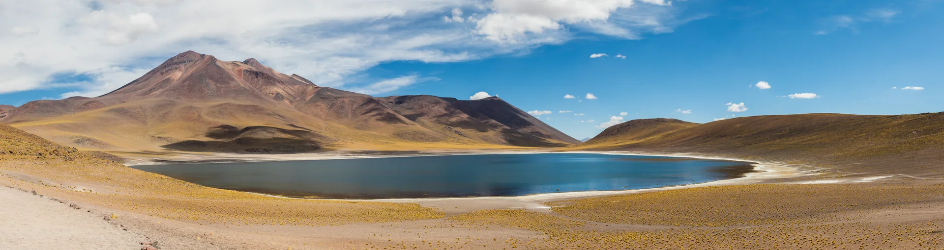 Panorama da Laguna Miñiques, de água azul escura entre montanhas áridas do altiplano sob céu com nuvens claras.