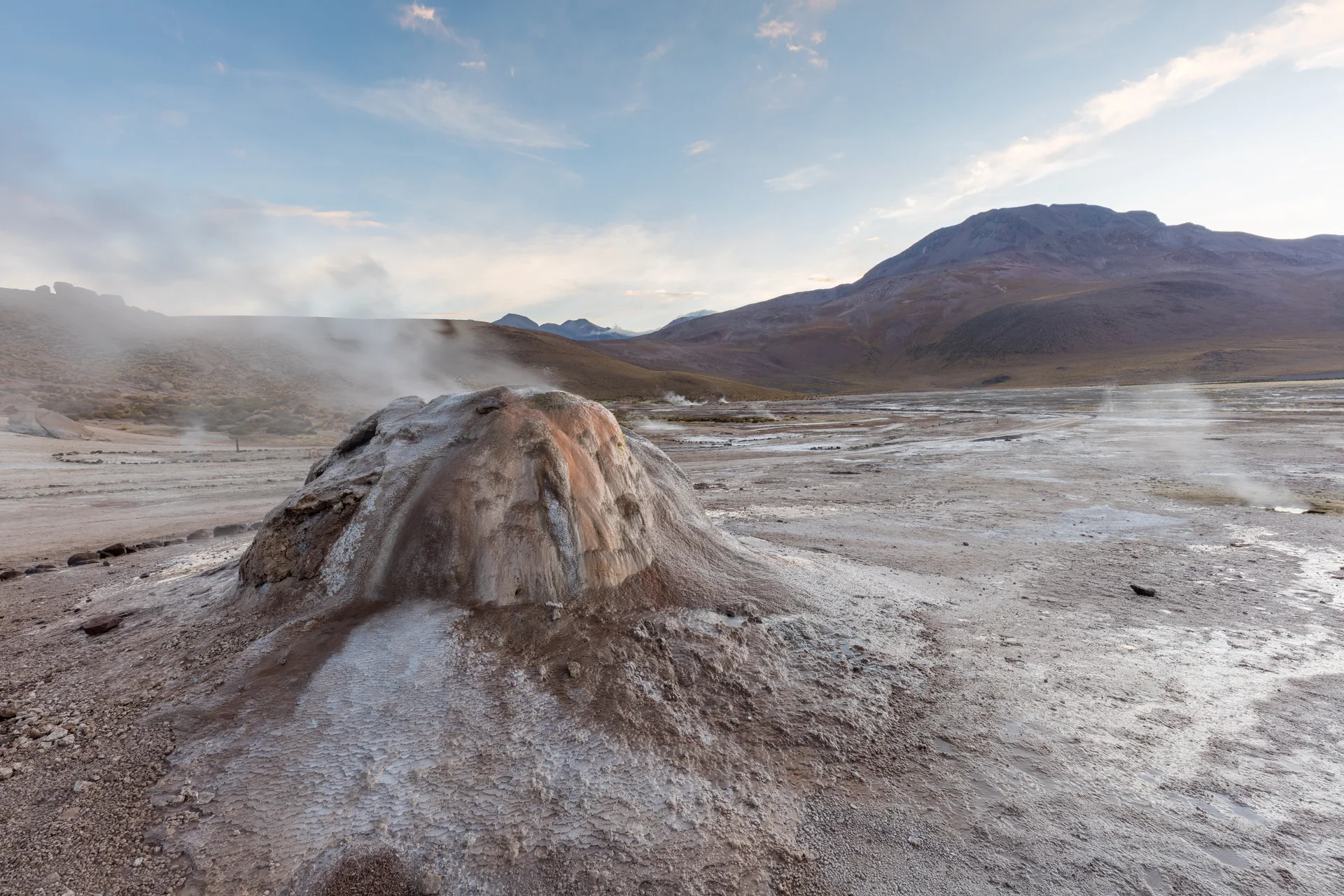 Vapor sobe de um cone geotérmico no campo de gêiseres de El Tatio, com montanhas do altiplano sob luz natural clara.