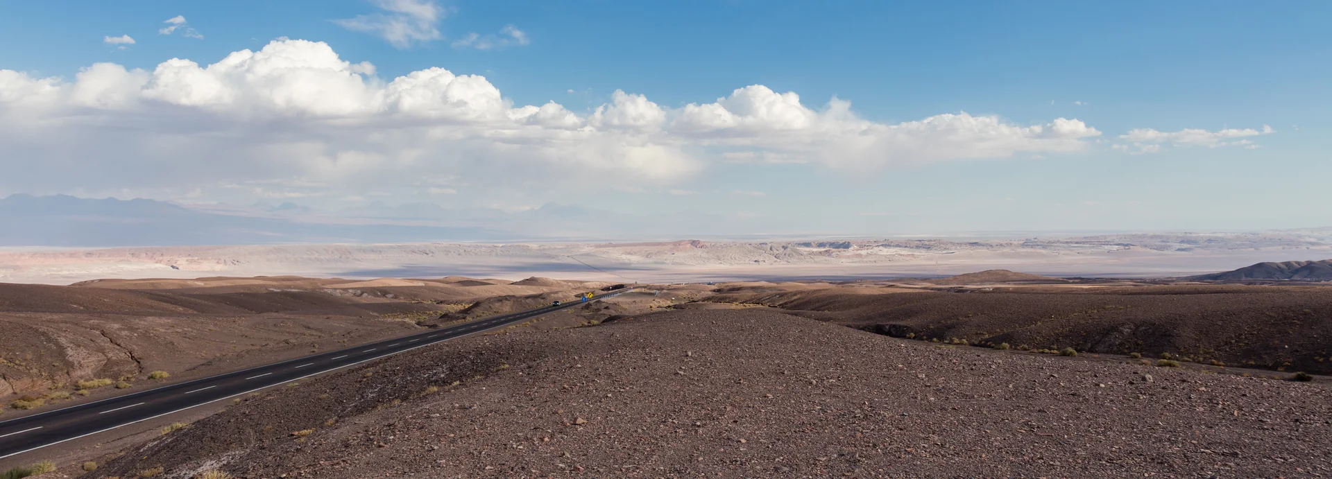 Estrada atravessa paisagem árida perto de San Pedro de Atacama, com planalto e montanhas ao fundo sob céu claro.
