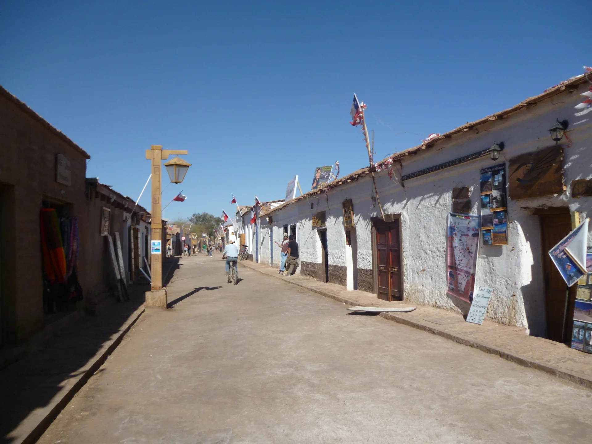 Rua de San Pedro de Atacama com fachadas baixas de adobe, pedestres e céu azul durante o dia.