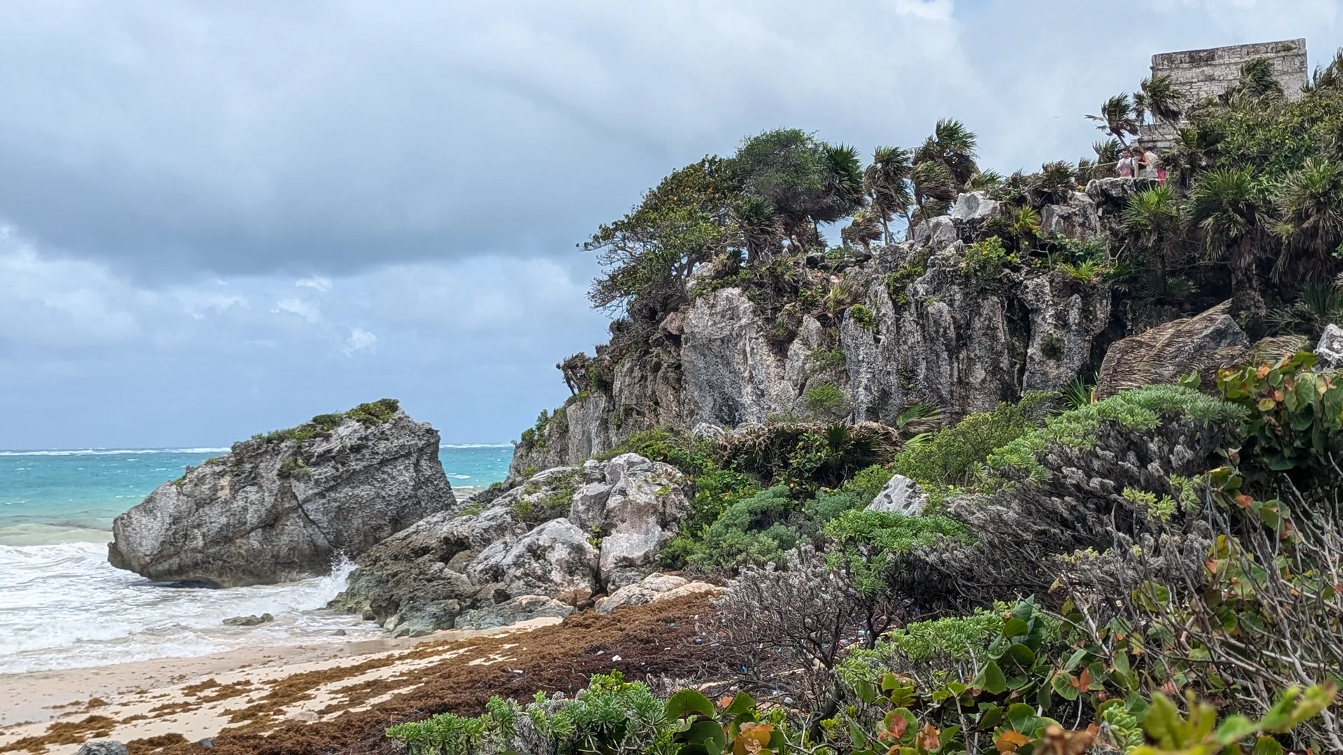 Litoral de Tulum durante o dia, com falésia calcária, vegetação, mar do Caribe e construção maia ao alto.