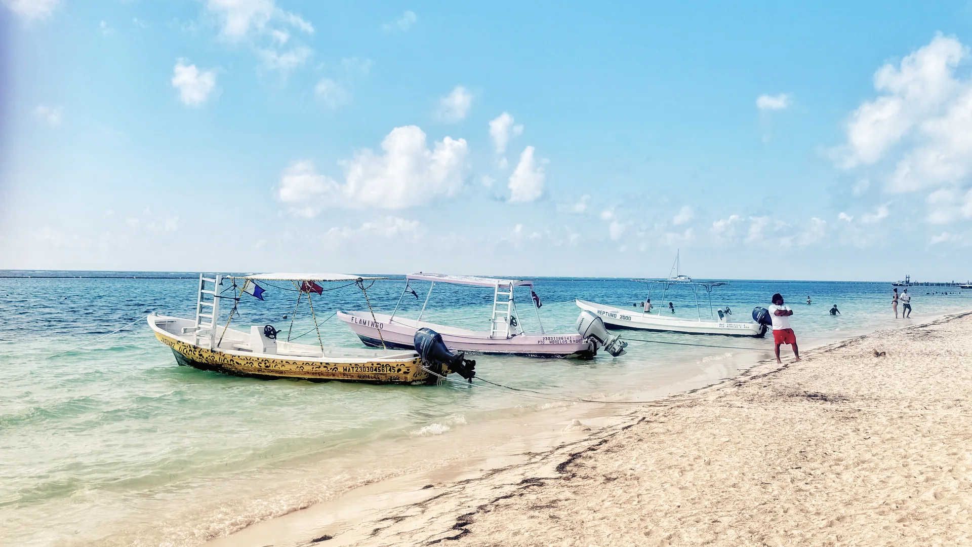 Praia de Puerto Morelos em dia claro, com pequenos barcos junto à areia, mar raso e píer ao fundo.