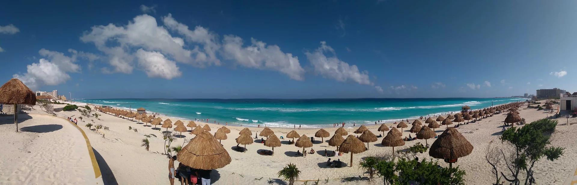 Panorama diurno da Playa Delfines, com faixa de areia, palapas, mar turquesa e céu parcialmente nublado em Cancún.