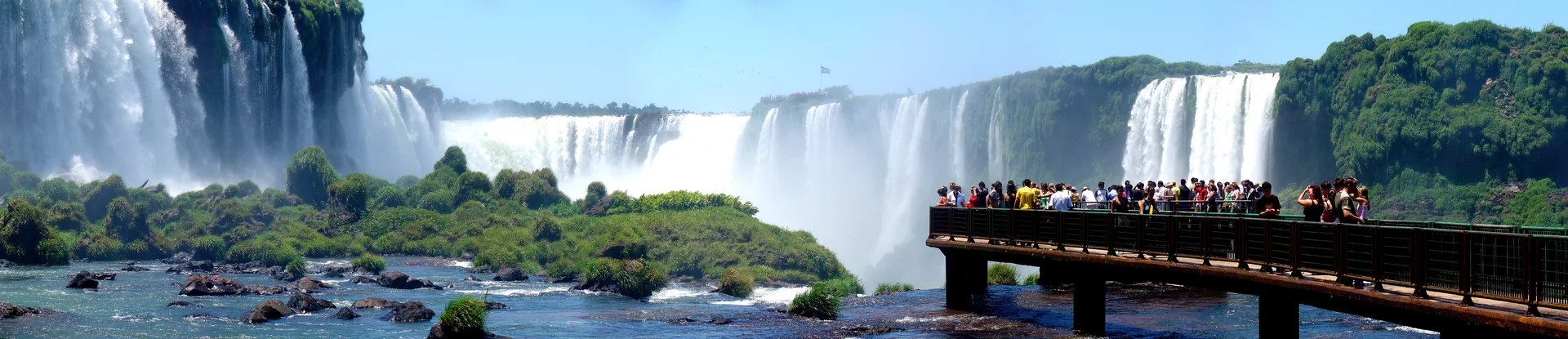 Panorama ensolarado das Cataratas do Iguaçu no Paraná, com quedas, vegetação e passarela do lado brasileiro.