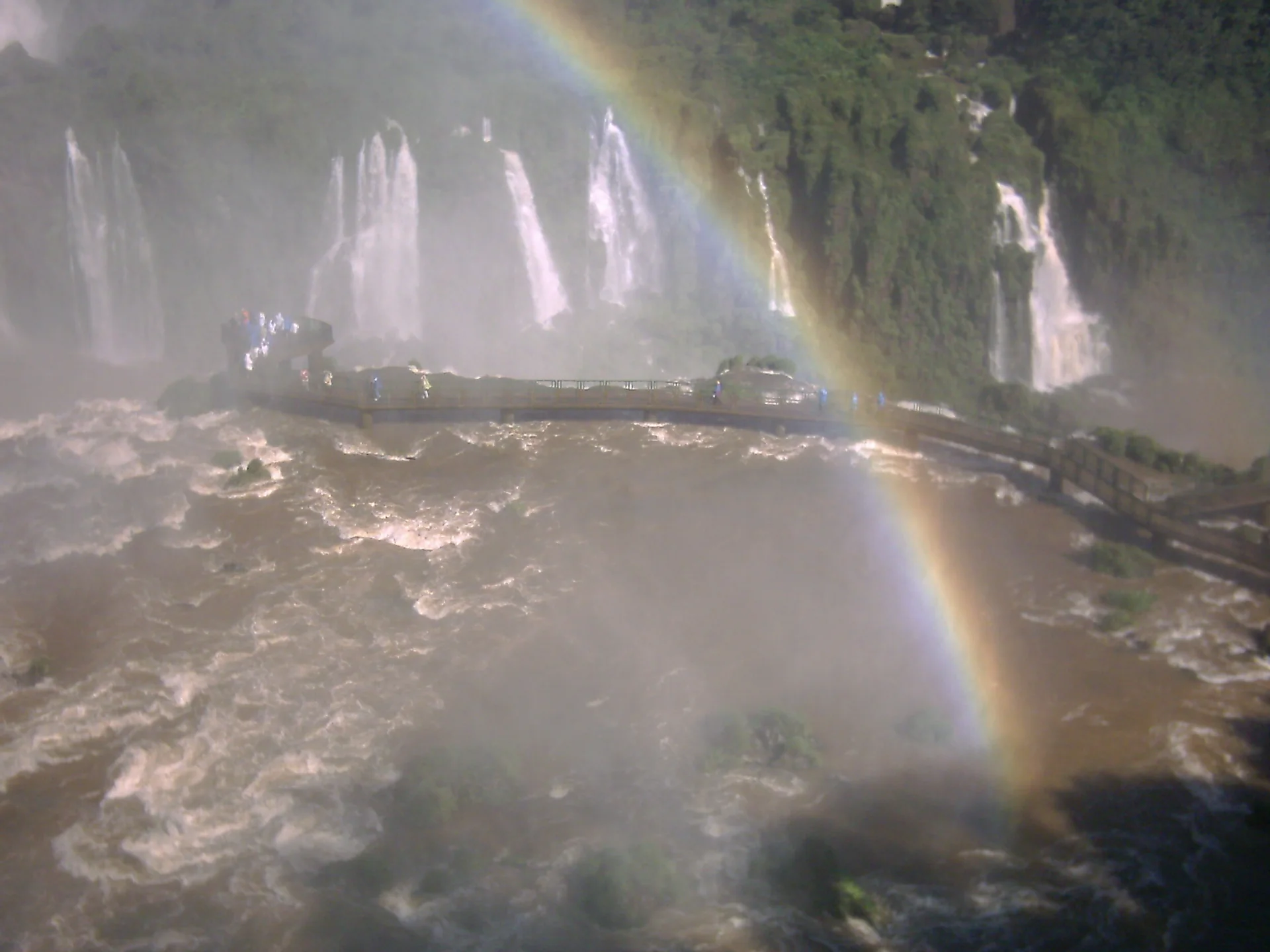 Vista elevada do lado brasileiro das Cataratas do Iguaçu, com arco-íris, passarela e quedas em meio à névoa.