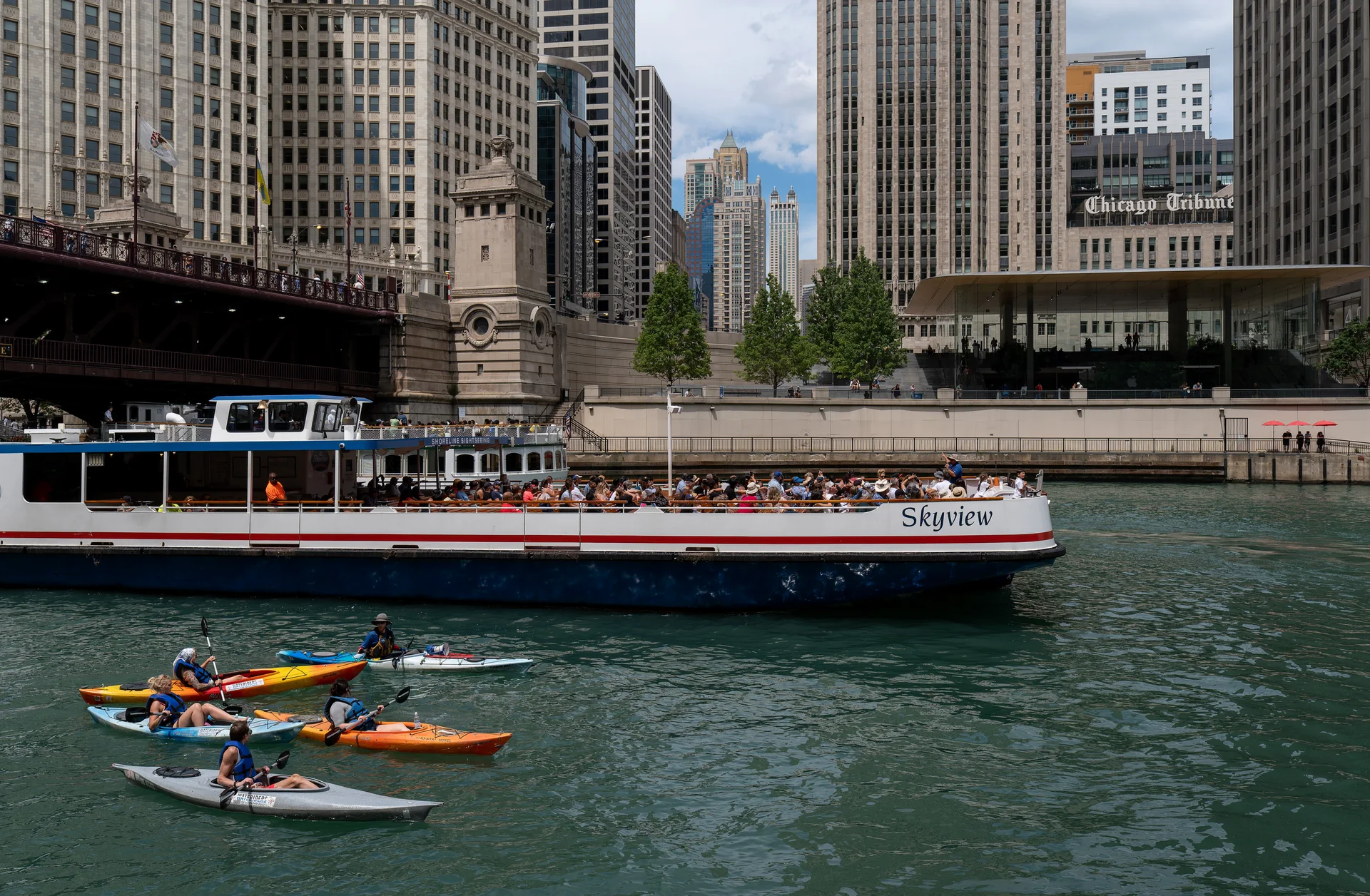 Caiaques coloridos e uma embarcação no rio Chicago, junto ao Riverwalk e a edifícios altos do centro em dia claro.