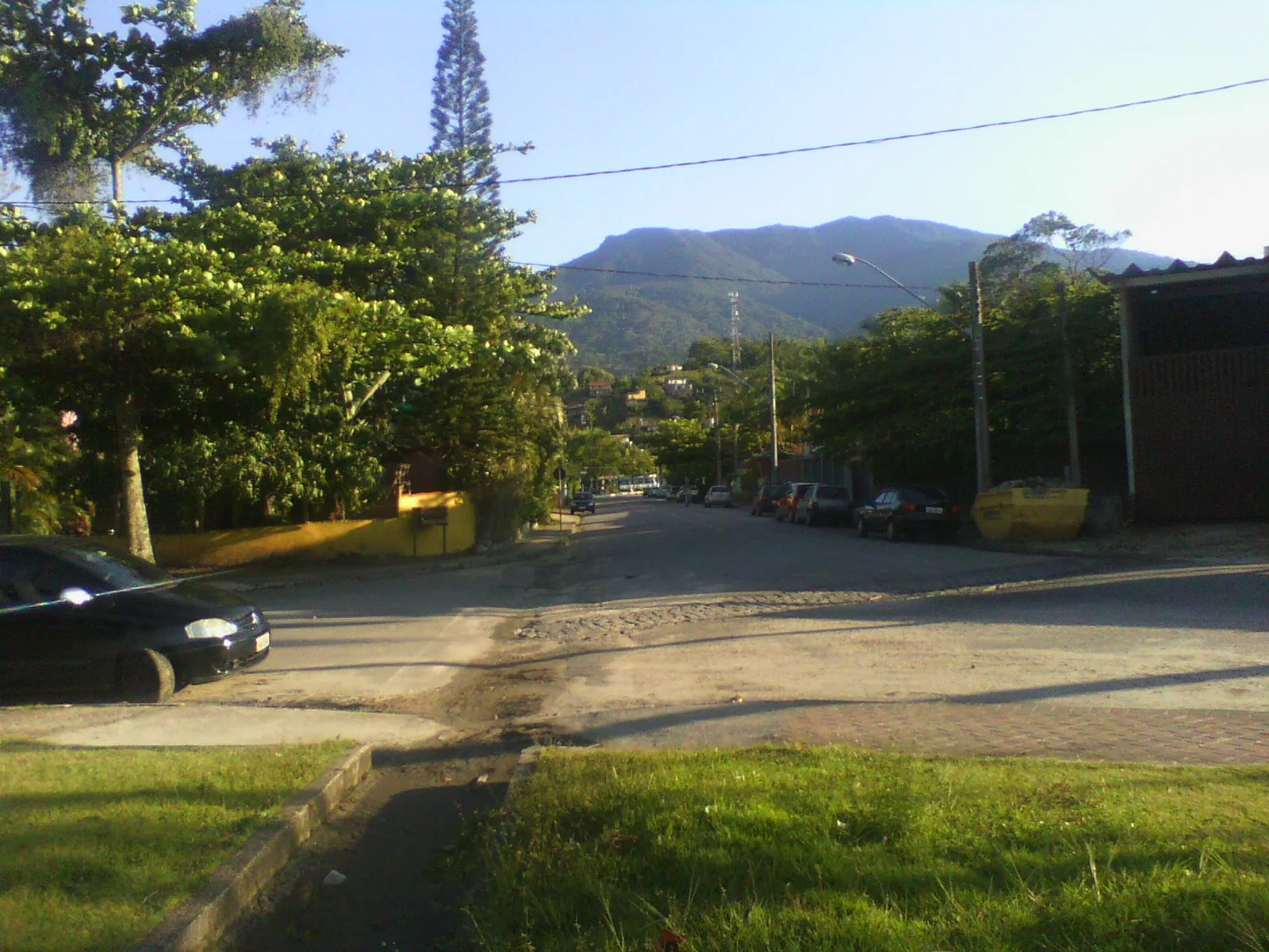 Via urbana arborizada em Ilhabela segue em direção ao relevo verde da ilha sob luz natural.
