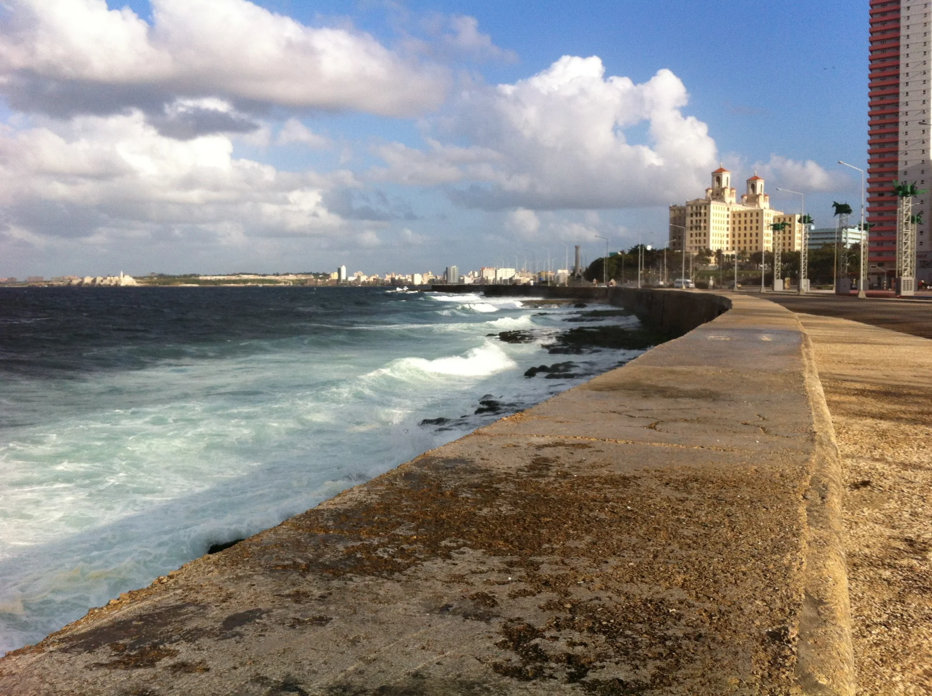 Calçadão do Malecón de Havana ao longo do mar, com prédios da cidade e céu parcialmente nublado durante o dia.