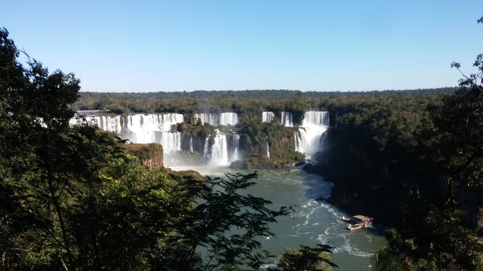 Vista ampla e ensolarada das Cataratas do Iguaçu e do Rio Iguaçu a partir da trilha no lado brasileiro.