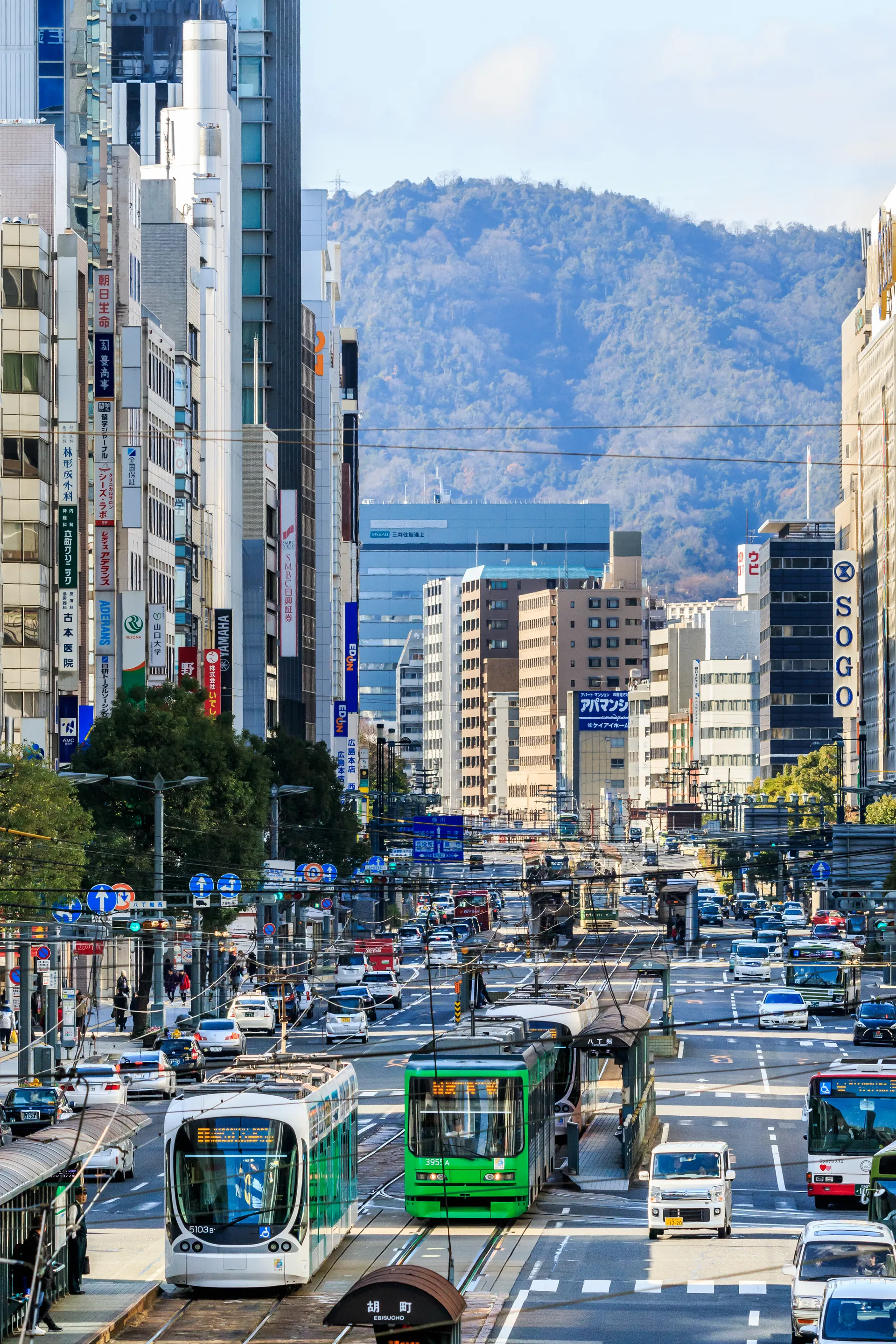Bondes verdes e brancos em corredor urbano do centro de Hiroshima, com morros ao fundo sob luz diurna.