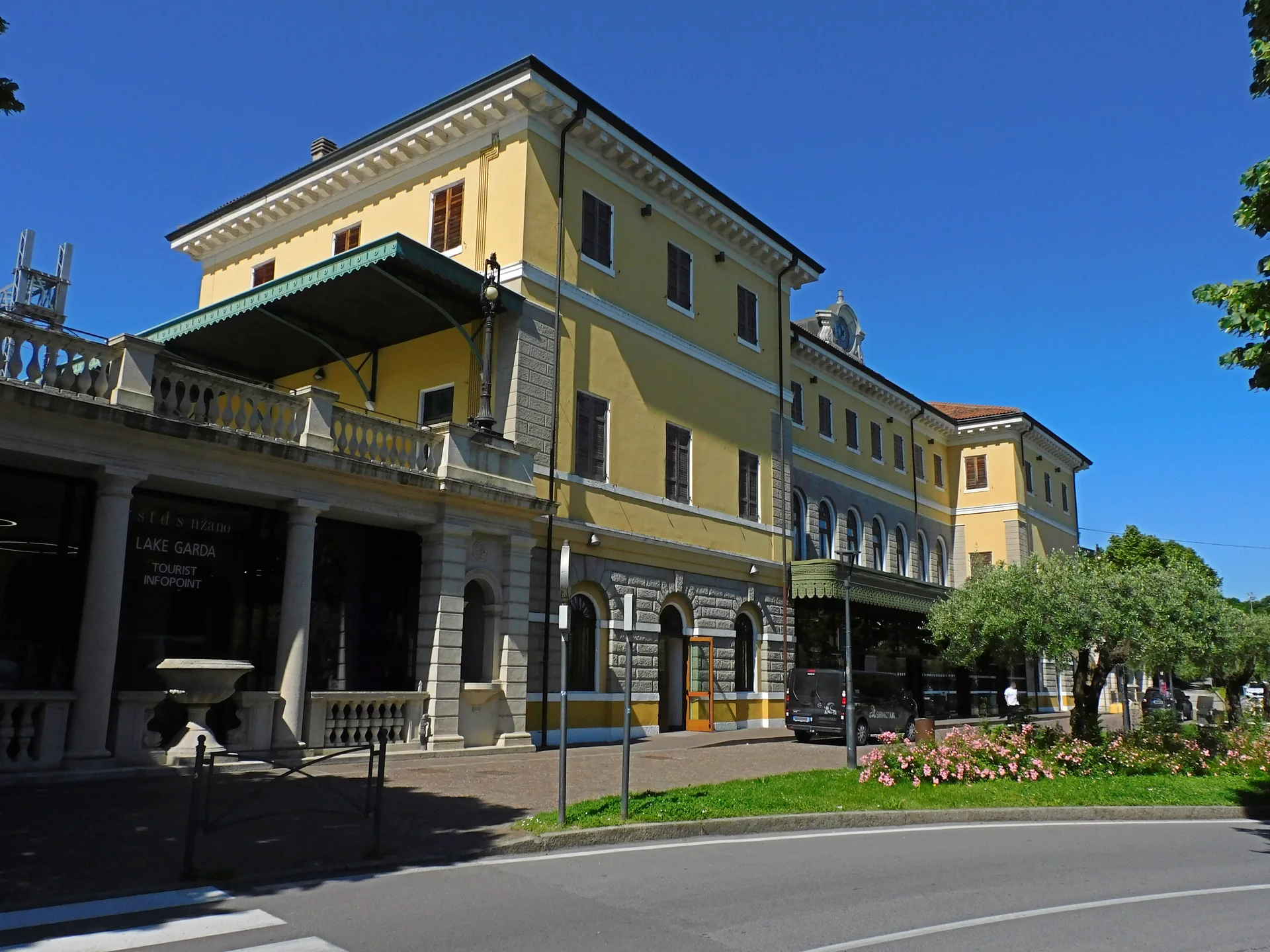 Fachada amarela da estação Desenzano del Garda–Sirmione sob céu azul, com árvores e via em primeiro plano.