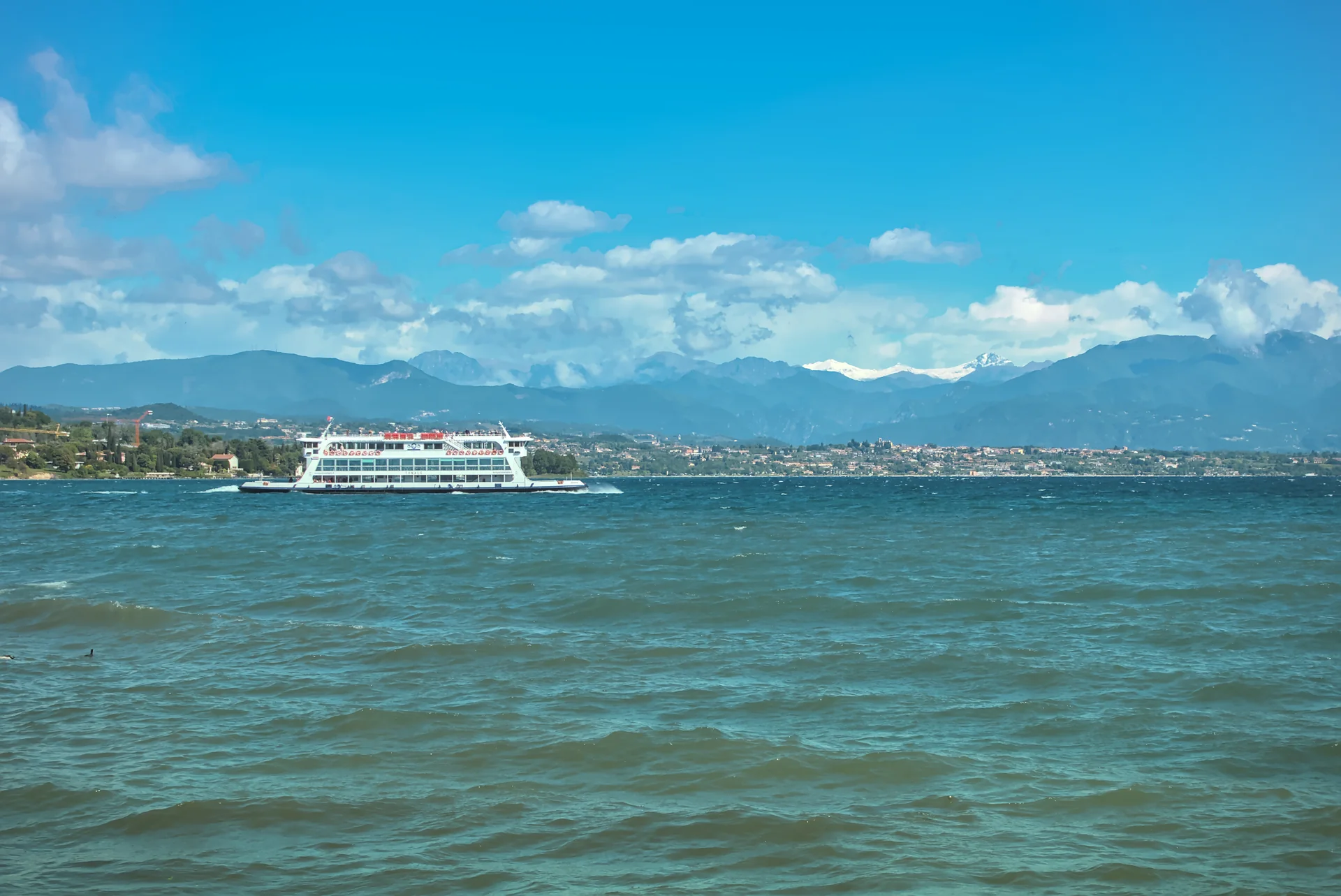 Balsa Tonale navegando após deixar Desenzano del Garda, com água azul-esverdeada e montanhas ao fundo.
