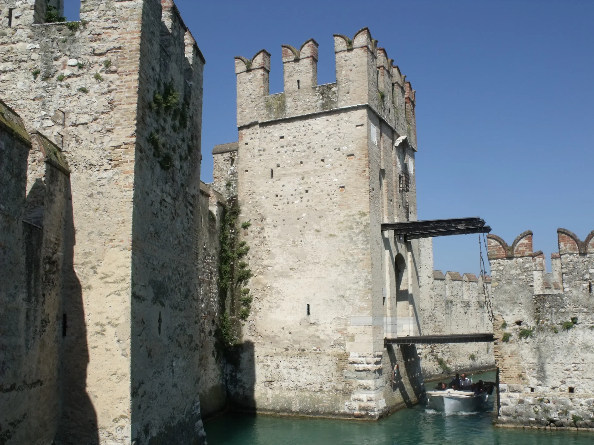 Ponte levadiça e muralhas do Castello Scaligero de Sirmione sob céu azul, com água do fosso em primeiro plano.