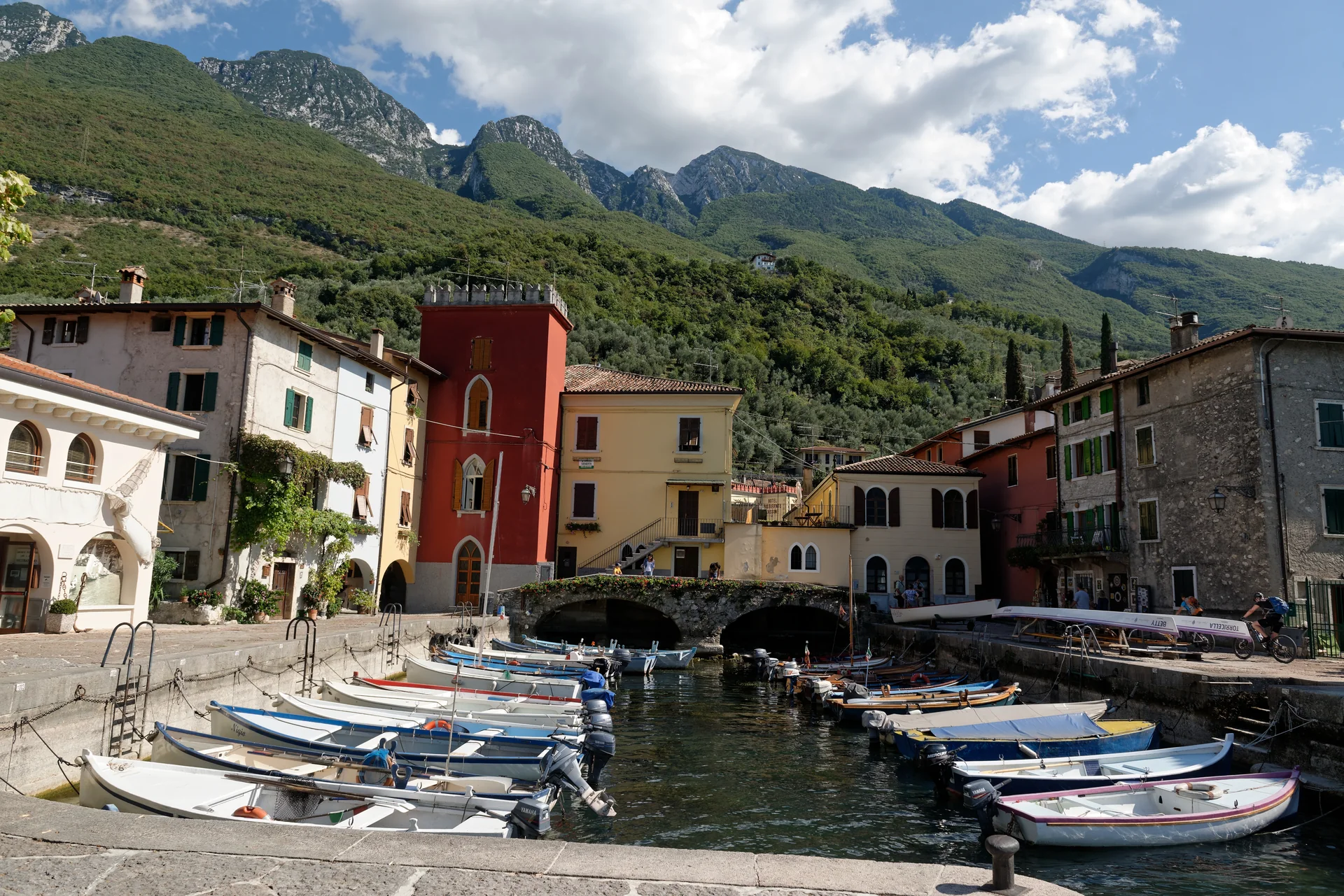 Pequeno porto de Cassone di Malcesine com barcos, casas coloridas e montanhas verdes sob luz natural.