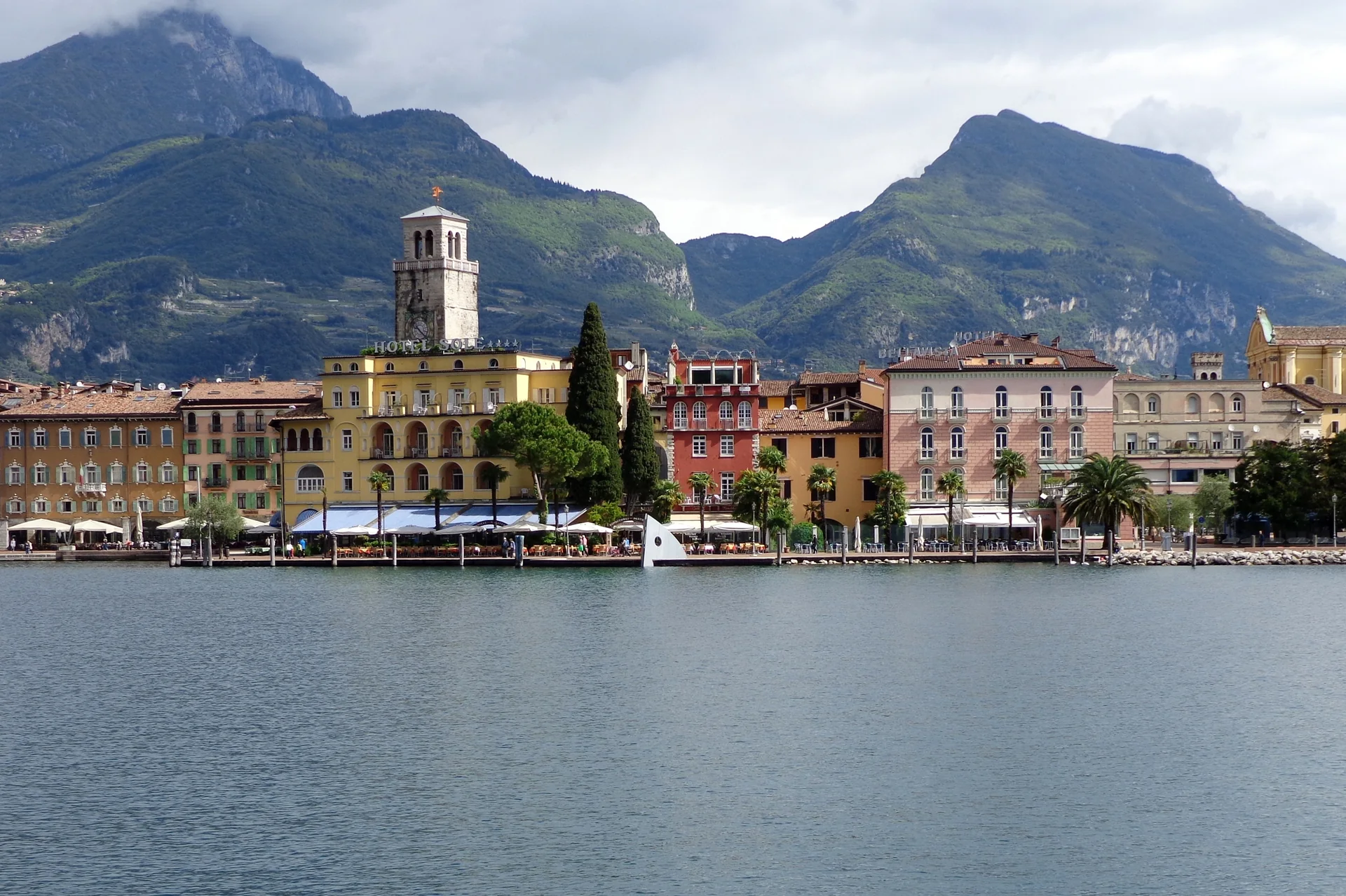 Orla de Riva del Garda vista do lago, com fachadas coloridas, torre, palmeiras e montanhas verdes ao fundo.