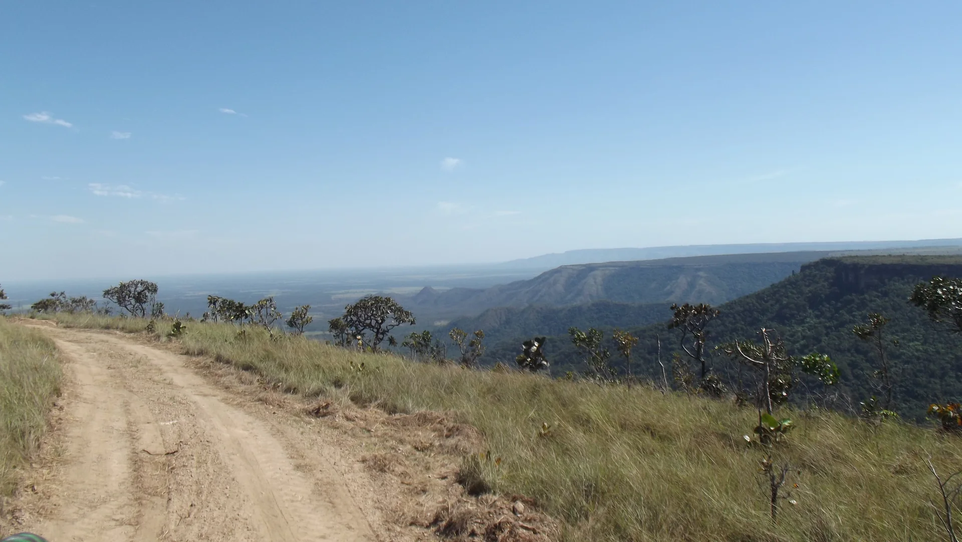 Estrada de terra na saída de uma trilha, com gramíneas do Cerrado e a paisagem da Chapada dos Guimarães ao fundo em dia claro.