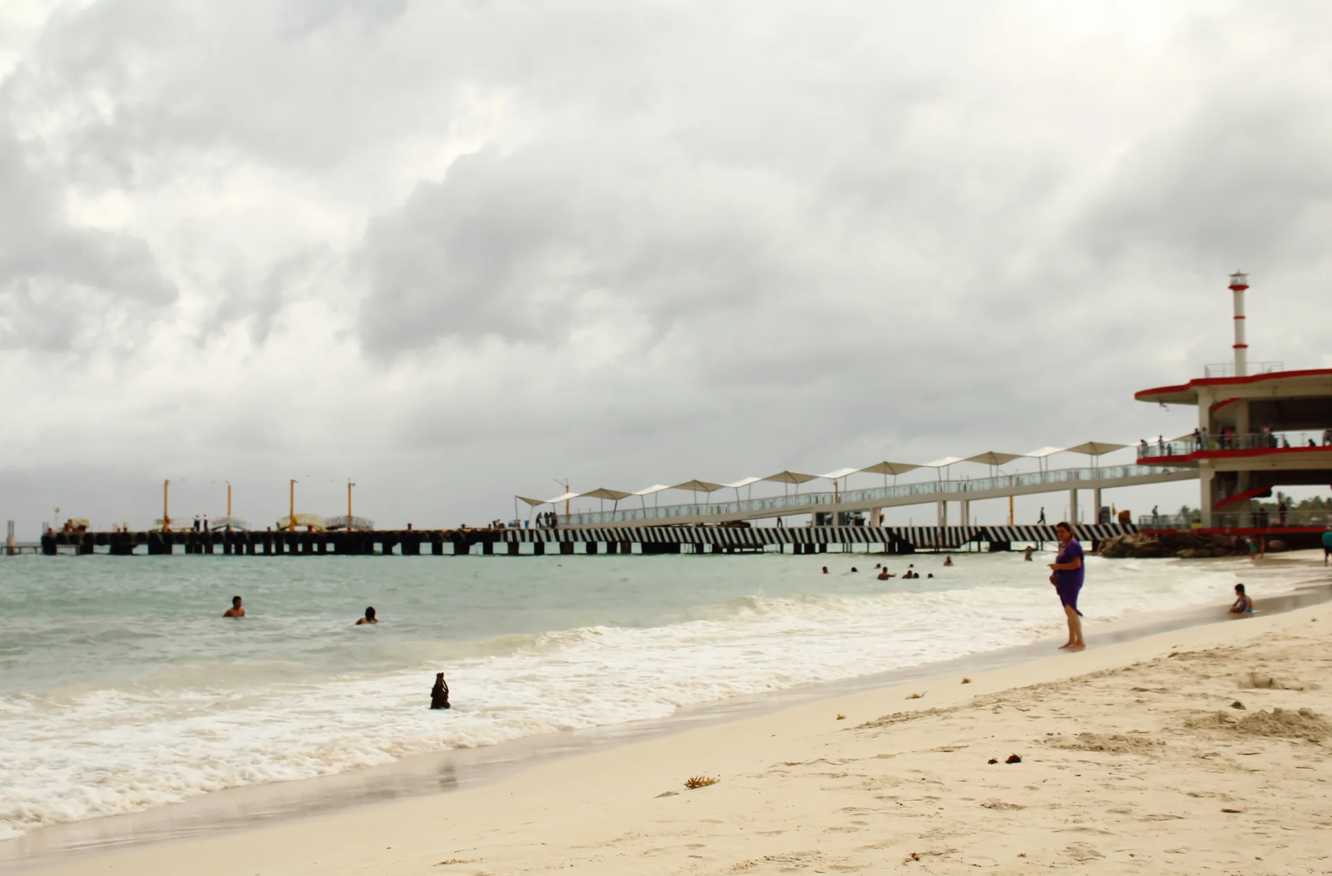 Praia junto ao píer de Playa del Carmen, com mar, faixa de areia e a estrutura do terminal sob luz diurna.