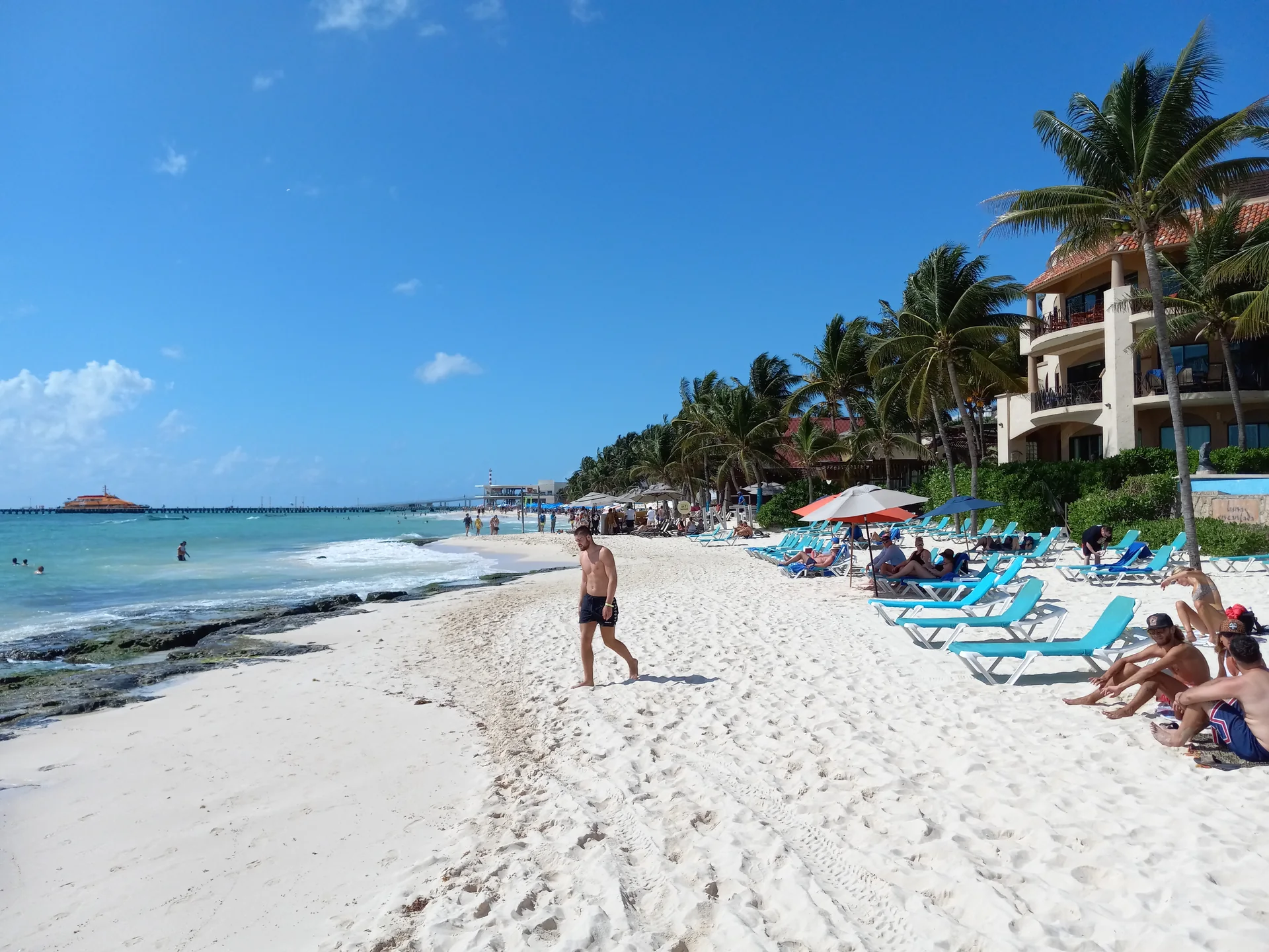 Faixa de areia de Playa del Carmen em dia claro, com mar Caribe, palmeiras e banhistas ao fundo.