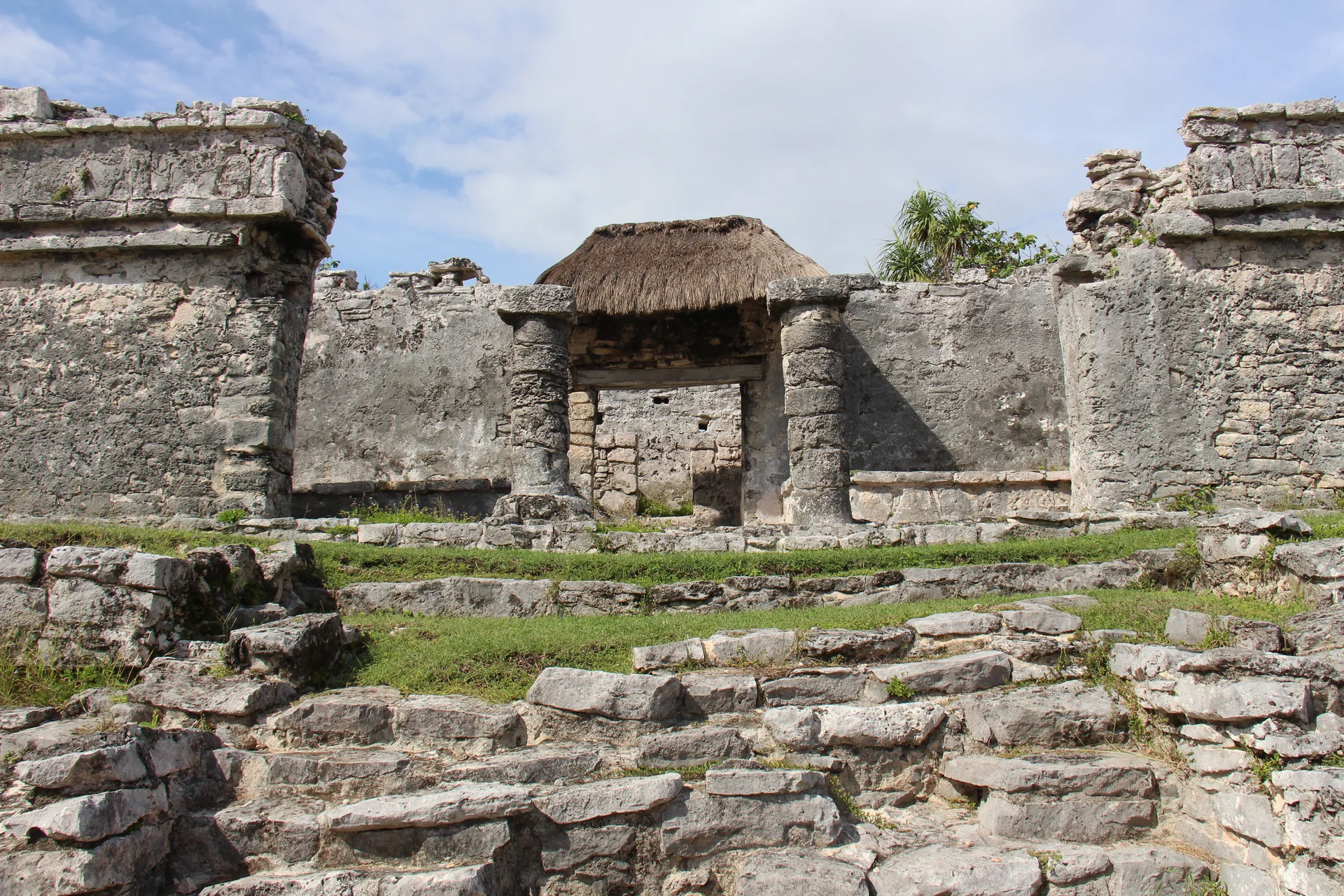 Estruturas de pedra das ruínas maias de Tulum sob luz natural, com vegetação e céu parcialmente nublado.