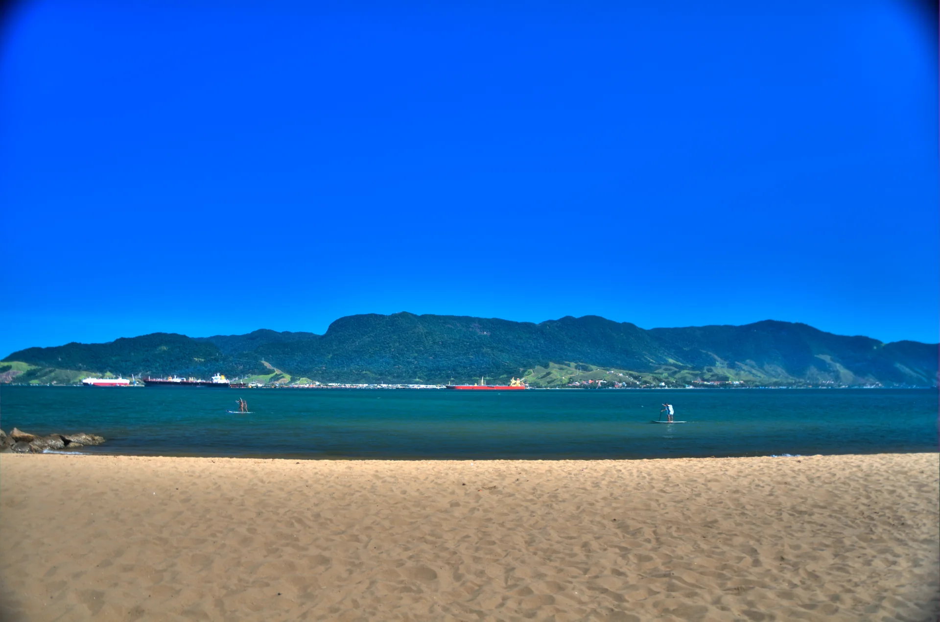 Faixa de areia da Praia do Perequê com mar do Canal de São Sebastião, embarcações ao longe e céu azul em Ilhabela.