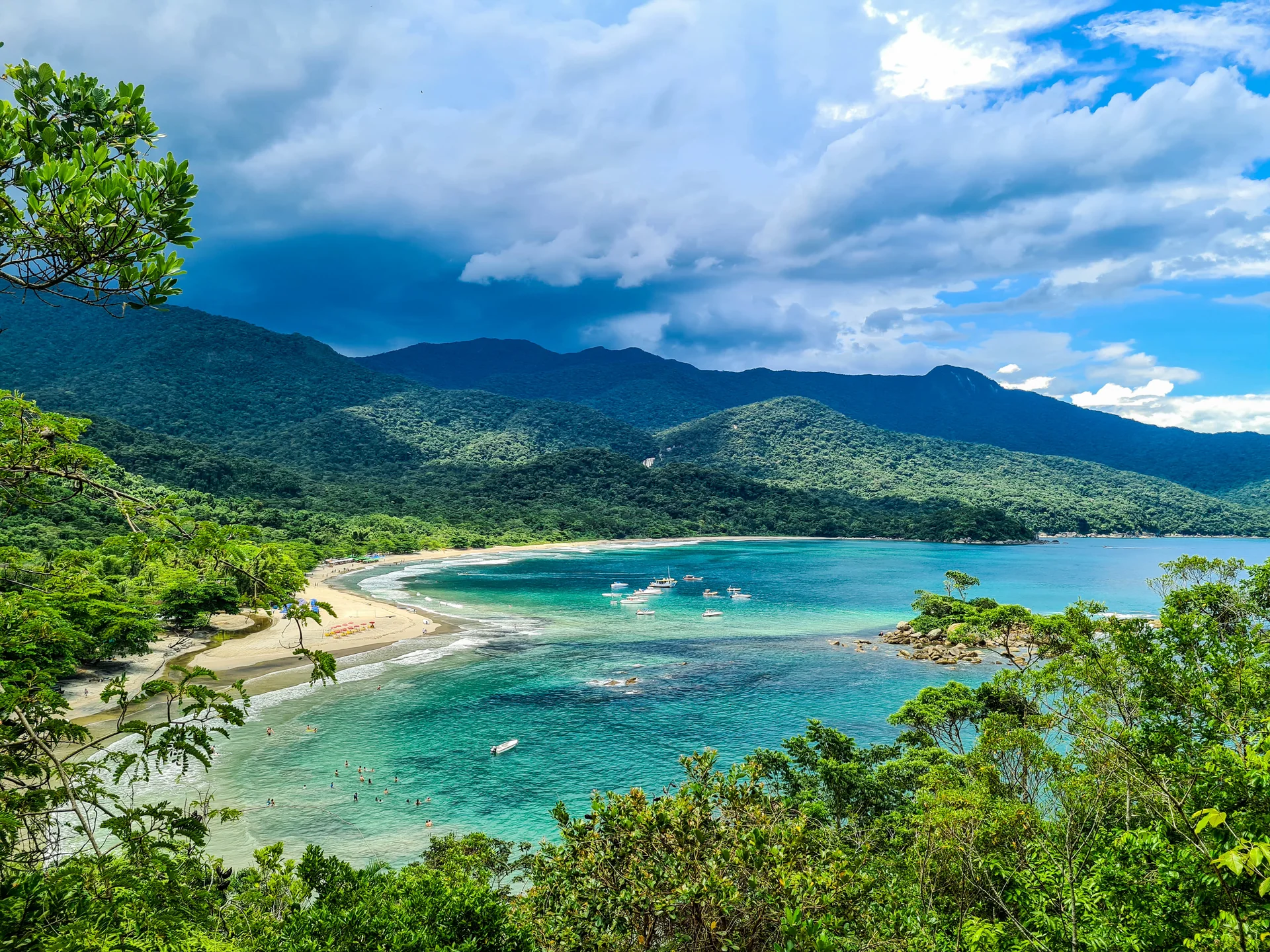 Vista elevada da Baía dos Castelhanos, com praia curva, mar azul-esverdeado, morros de Mata Atlântica e céu parcialmente claro.