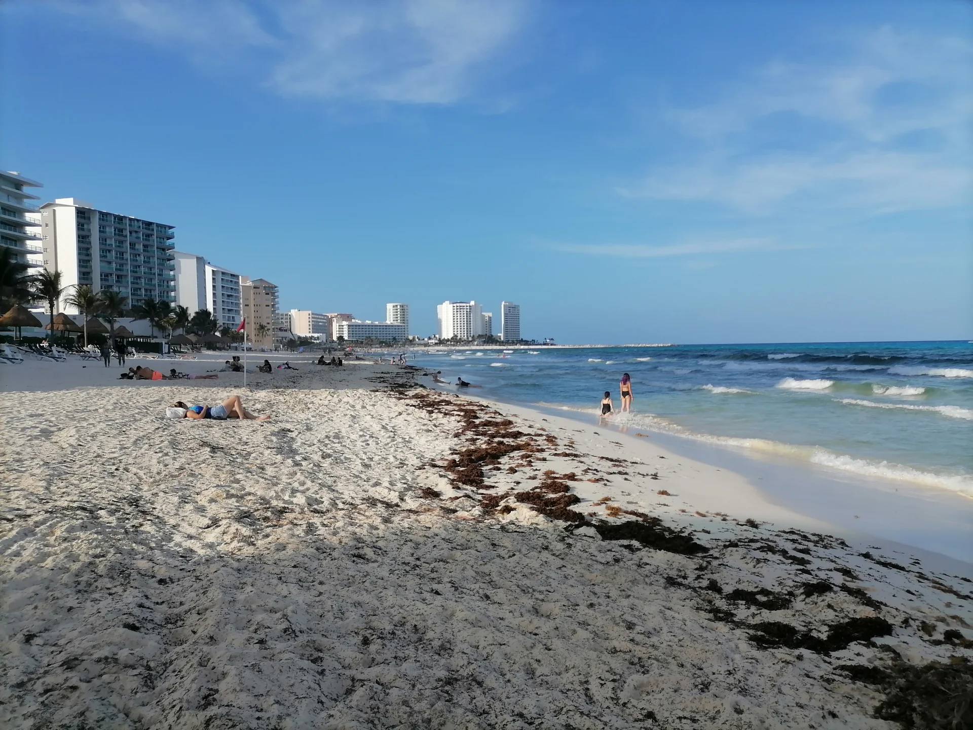 Playa Delfines em Cancún durante o dia, com areia, mar, edifícios ao fundo e uma faixa de sargaço visível na praia.