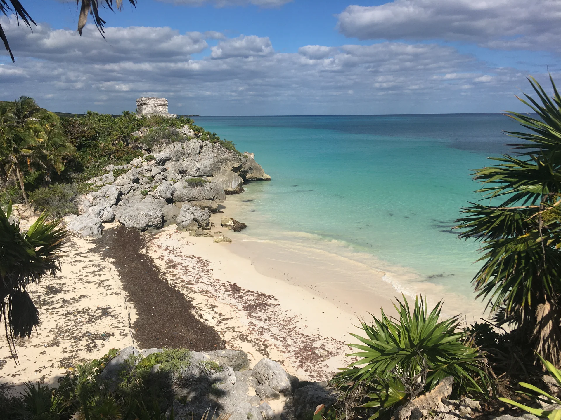 Ruínas maias de Tulum sobre uma falésia, emolduradas por vegetação, praia e mar do Caribe sob luz diurna.