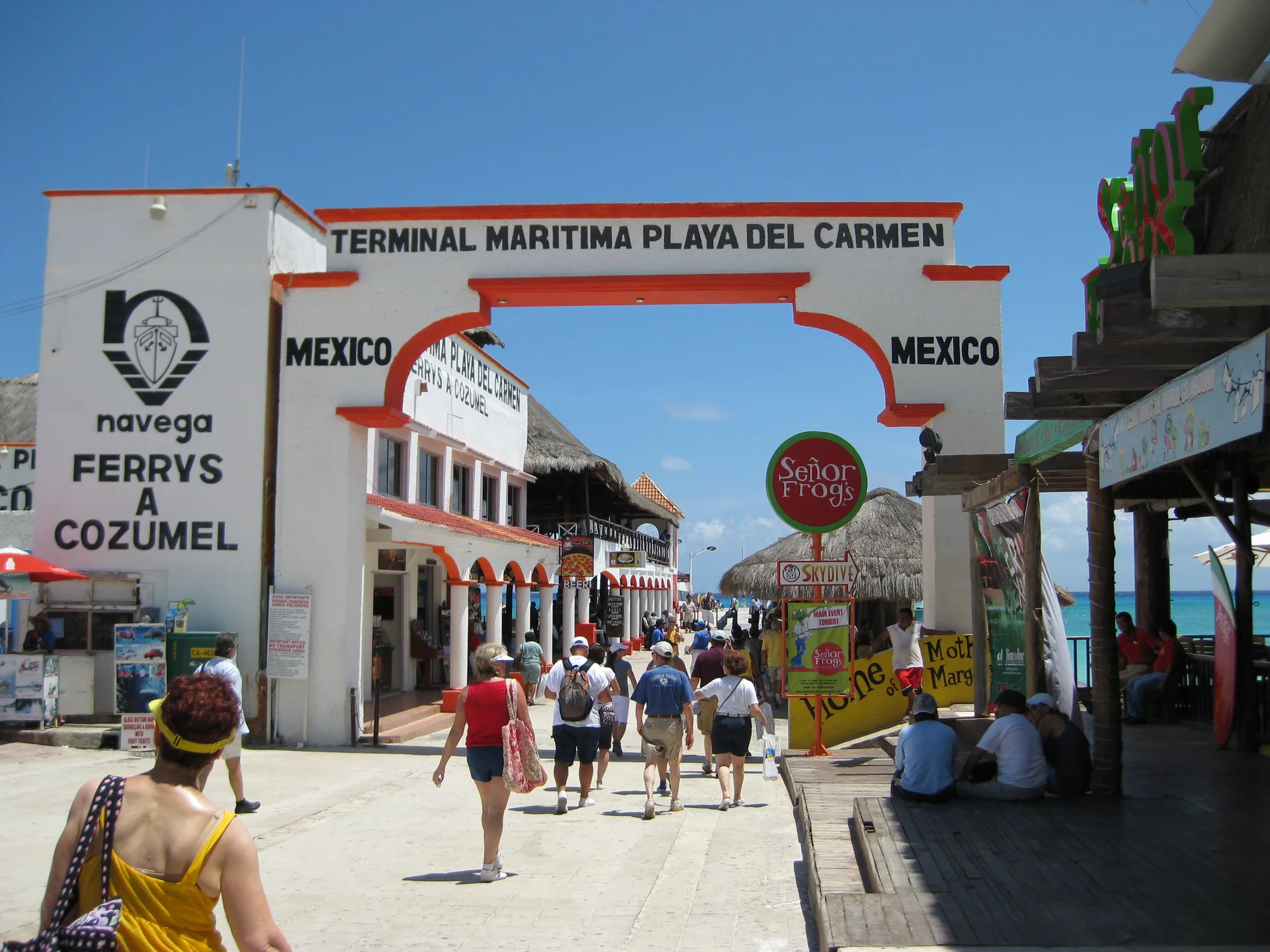 Entrada do Terminal Marítimo de Playa del Carmen em dia ensolarado, com viajantes caminhando em direção ao acesso de ferries para Cozumel.