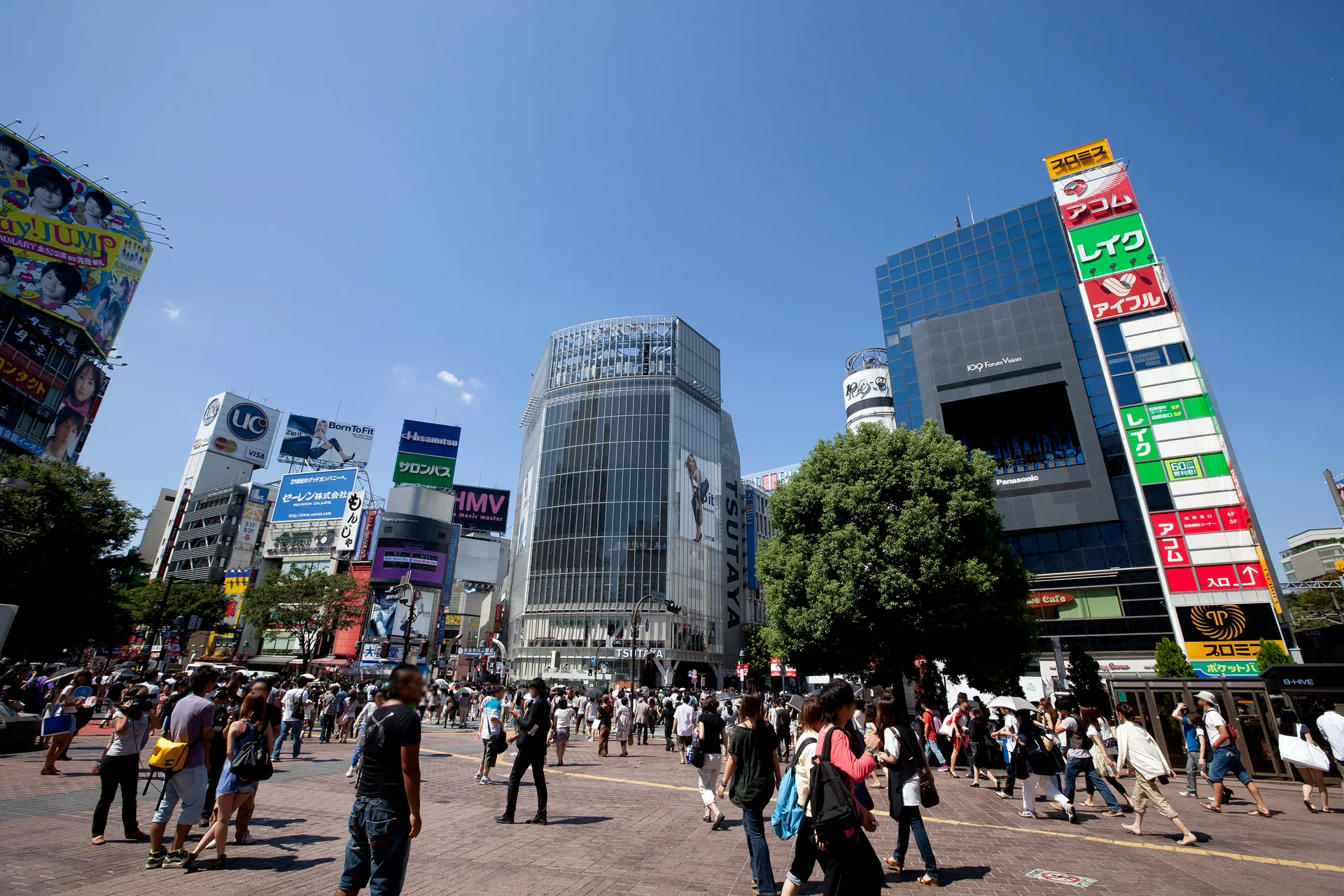 Entorno da Estação Shibuya em dia ensolarado, com calçada, pedestres e edifícios comerciais.