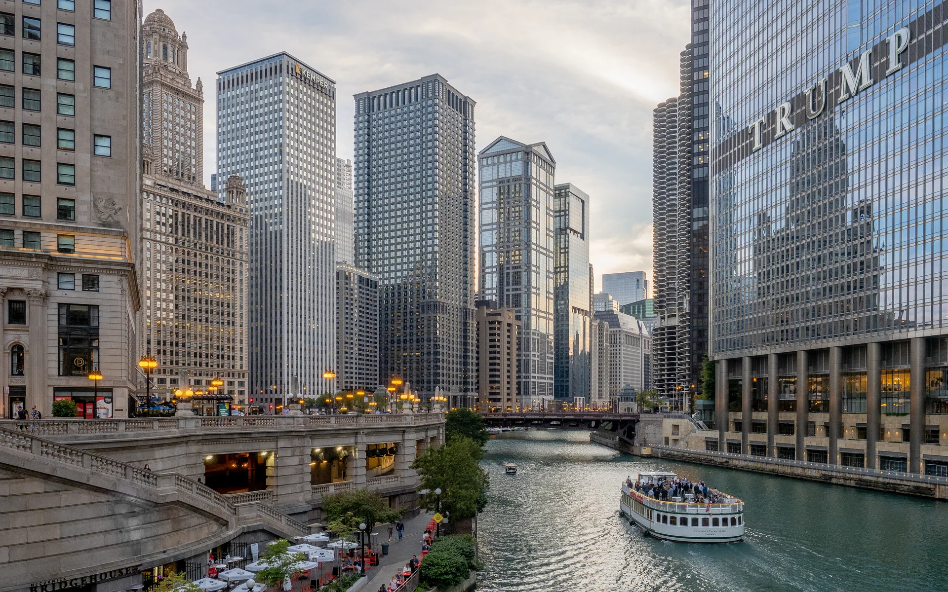 Vista elevada do Chicago Riverwalk e do rio, com embarcação, pontes e torres do centro em luz de fim de tarde.